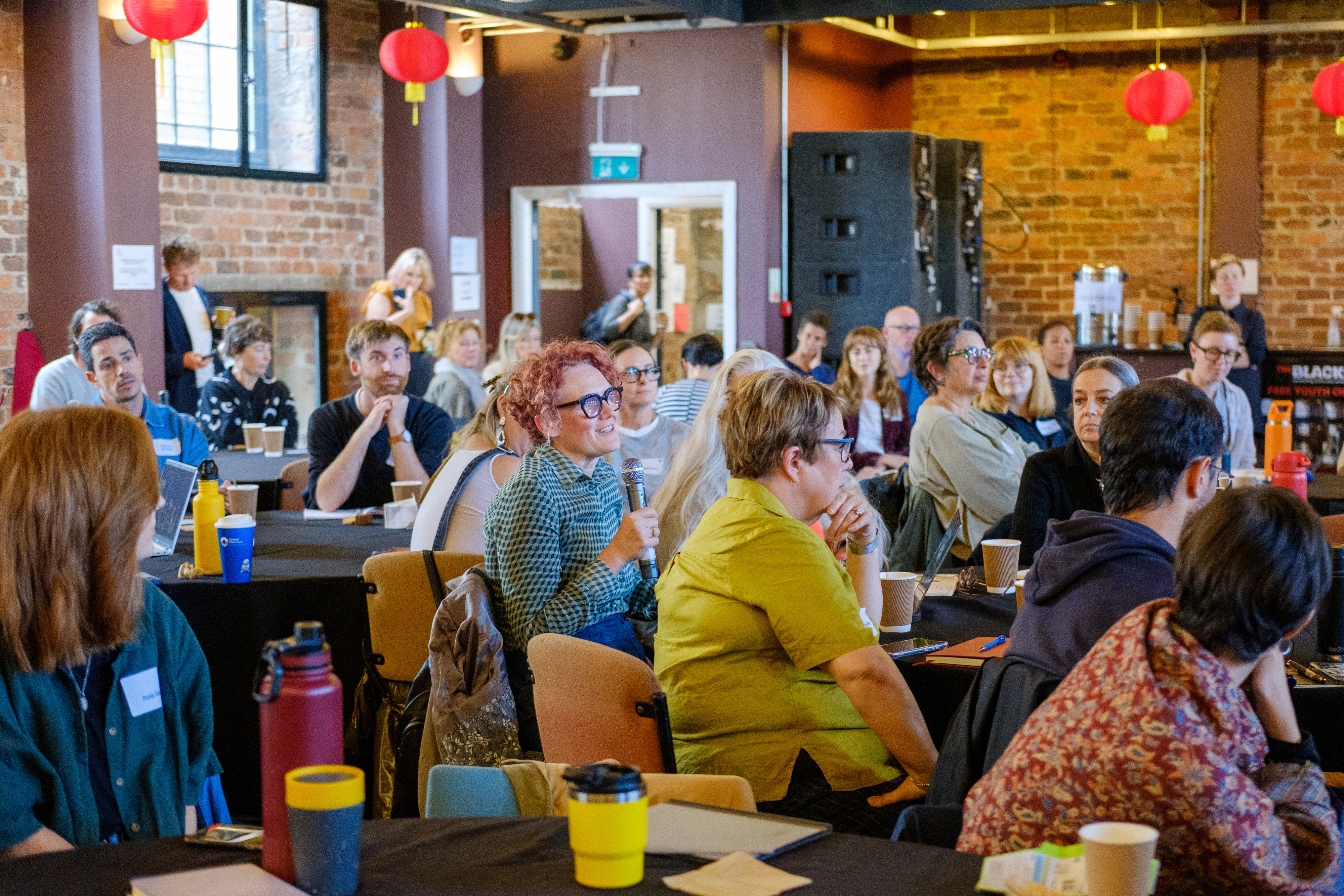 A large room full of people sitting around big circular tables.