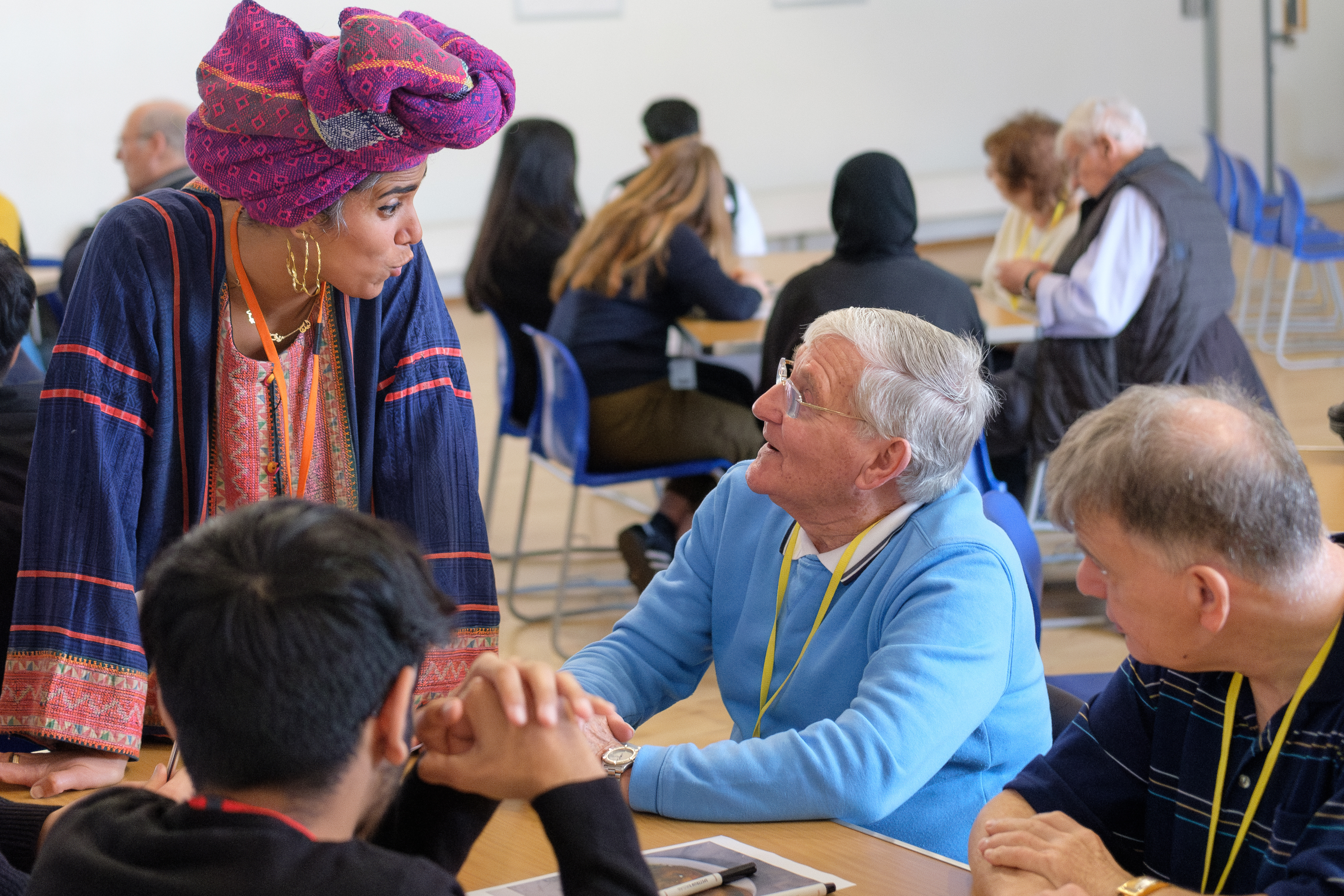 A group of elderly people sitting around a table wearing yellow lanyards. One person is looking up at a woman speaking to them standing up. The room is full of other people sitting around tables.