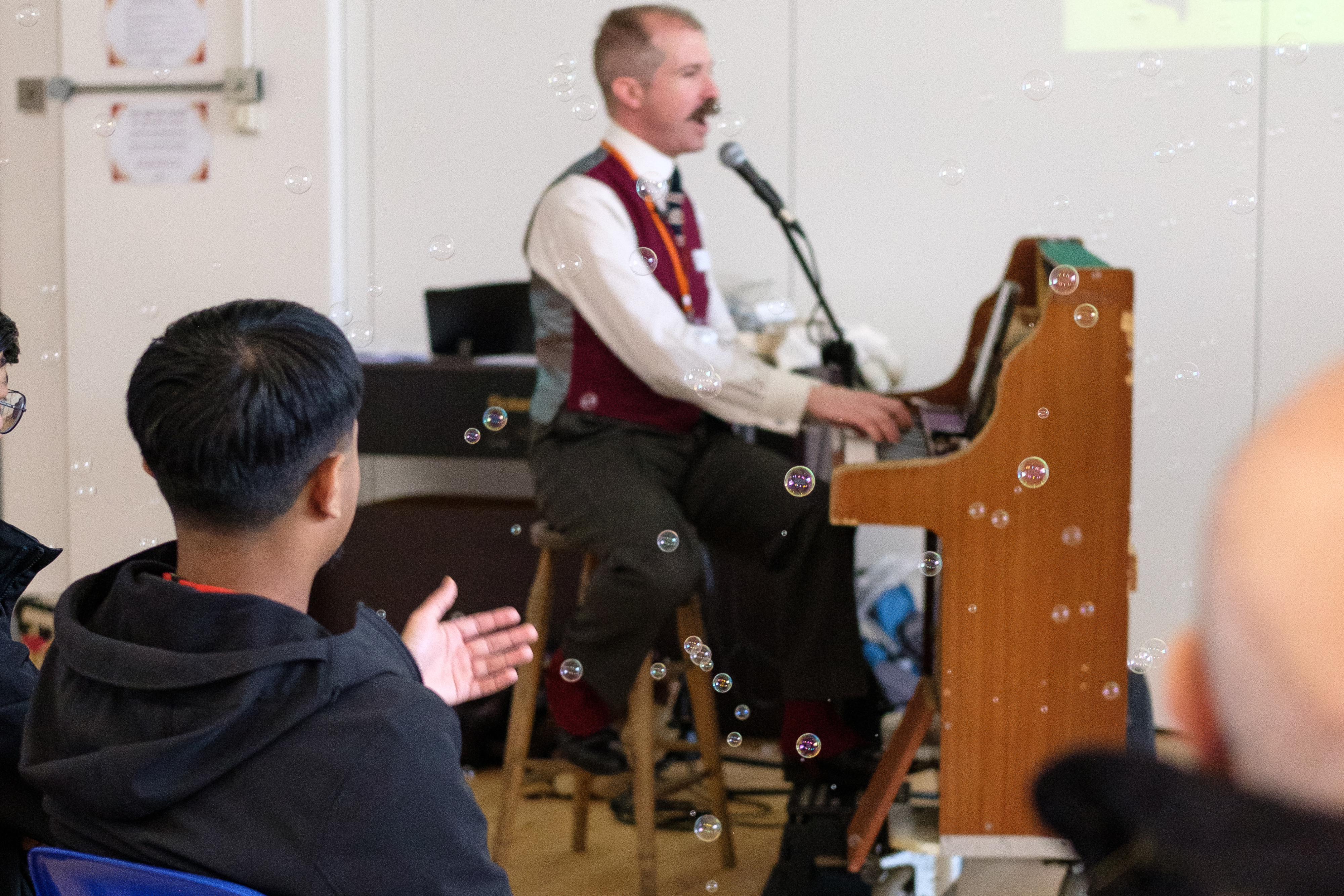A teenage boy reaches out towards bubbles. Behind the bubbles, A person is playing the piano who is wearing a waistcoat and has a moustache.