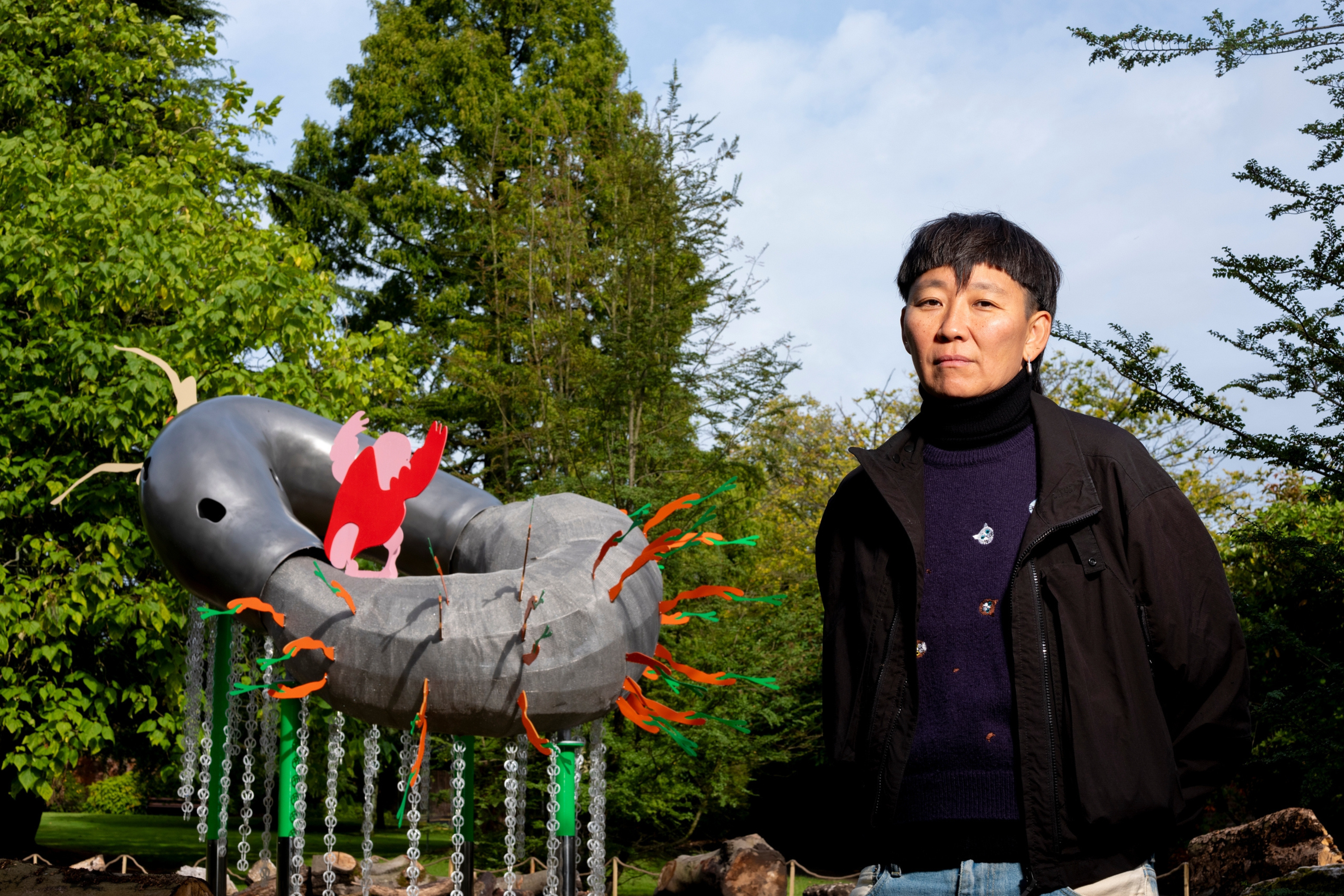 Anne Duk Hee standing in front of a large metal sculpture standing above a pond. The sculpture has green and orange snake-like bits poking out, and a large pink and red creature on top.