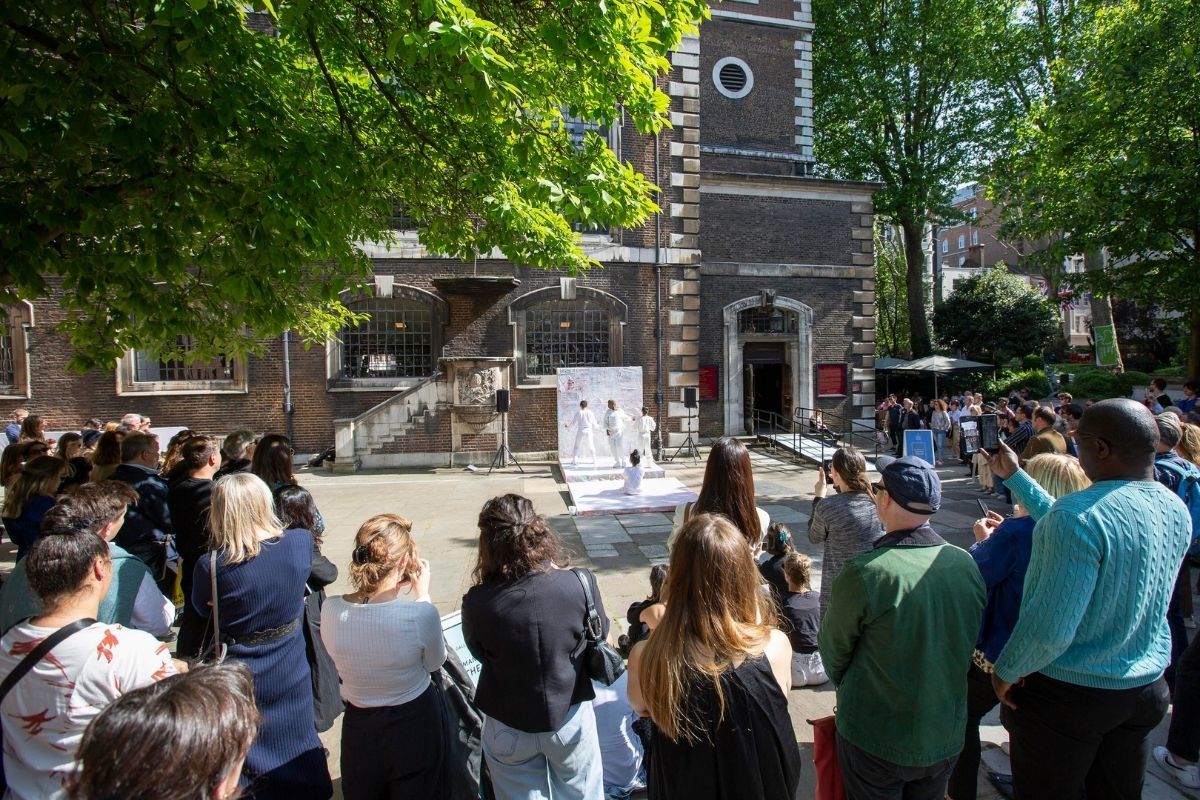 Three dancers in white outfits dancing in a line, leaning backwards on a stage, with another dancing sitting down on a stage and watching them, in front of a large audience.