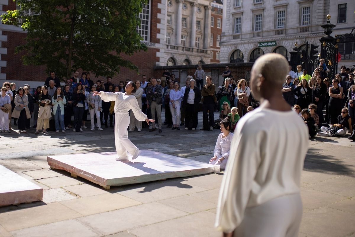 A dancer in a white outfit on a stage, with another dancer watching them, in front of a large audience.