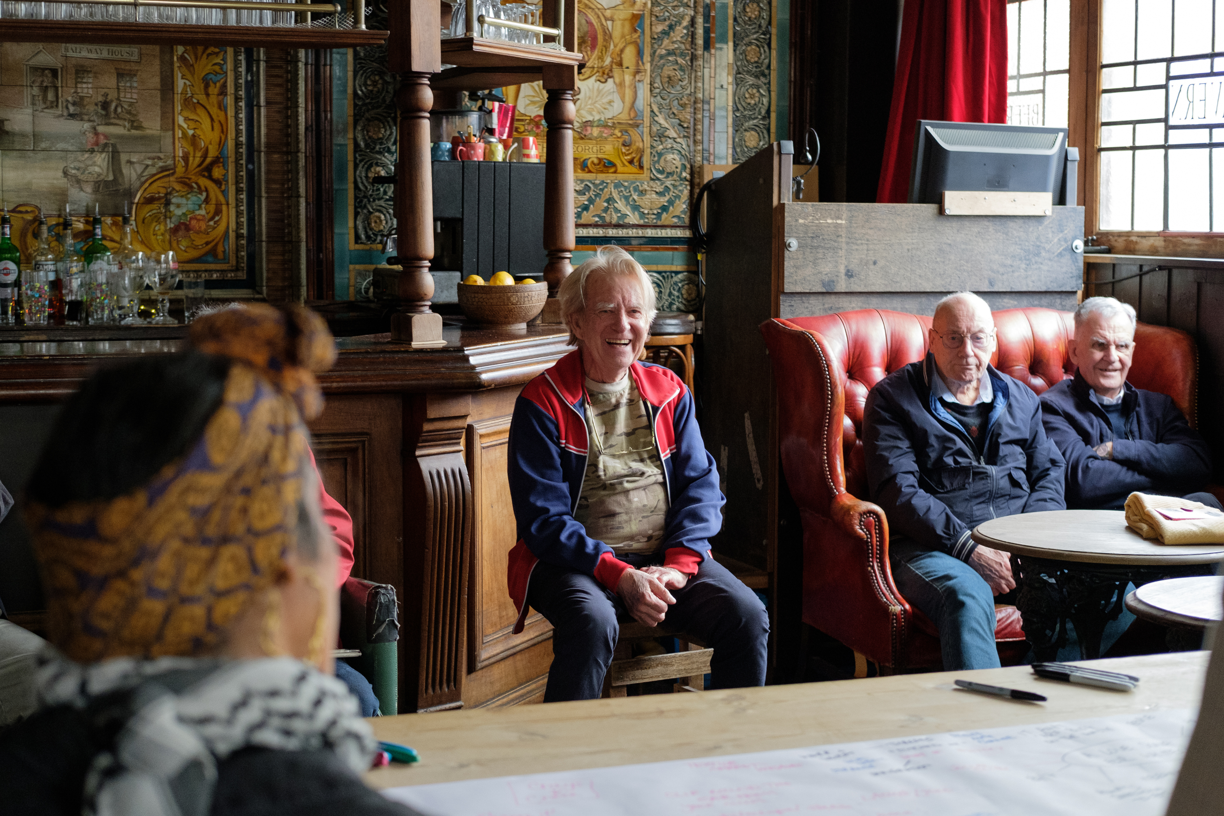 A group of elderly men sitting around in an old-style pub with dark wooden benches and big windows.