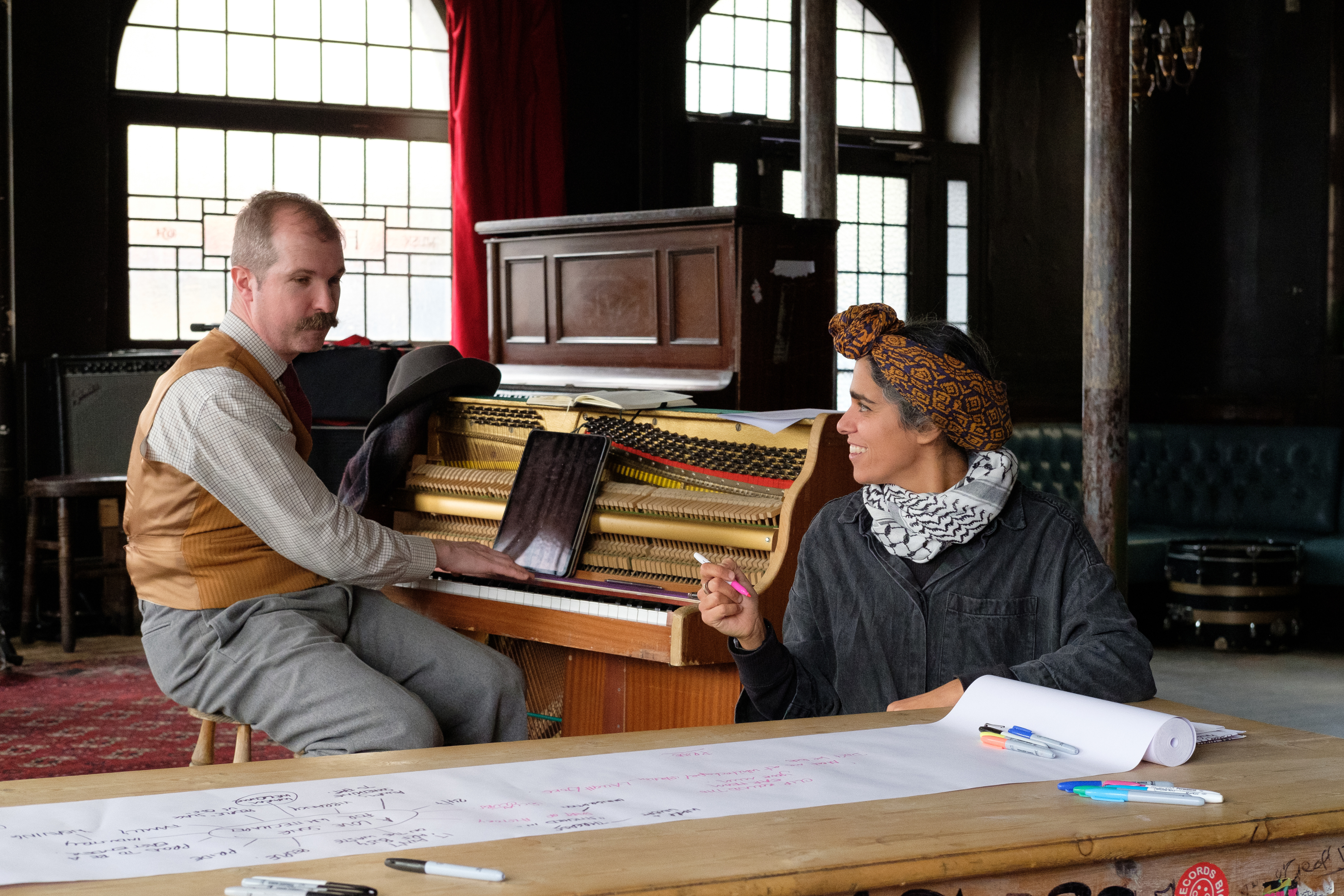 Two people sitting in a large room with big windows and red curtains. One is sitting by a piano and the other is writing on a long roll of paper on large table.