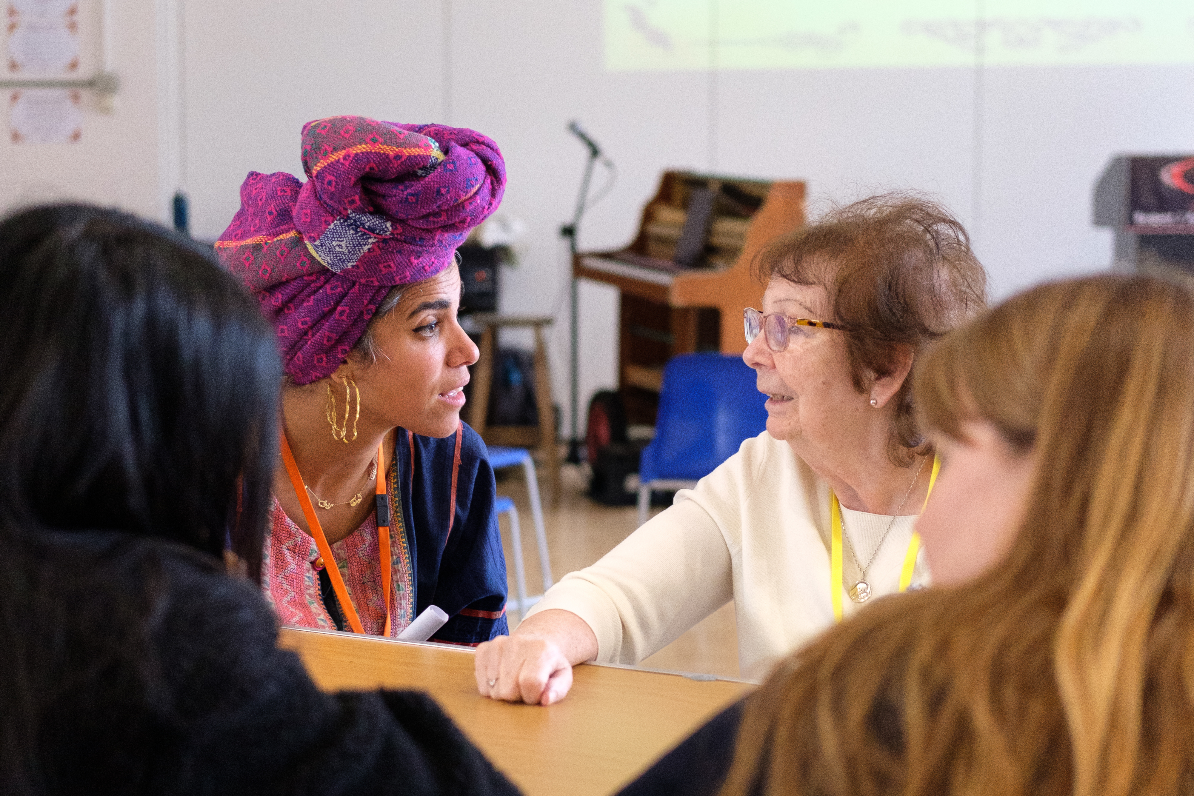 An elderly woman is looking at another woman both speaking. They are joined by two people facing them around a wooden table.