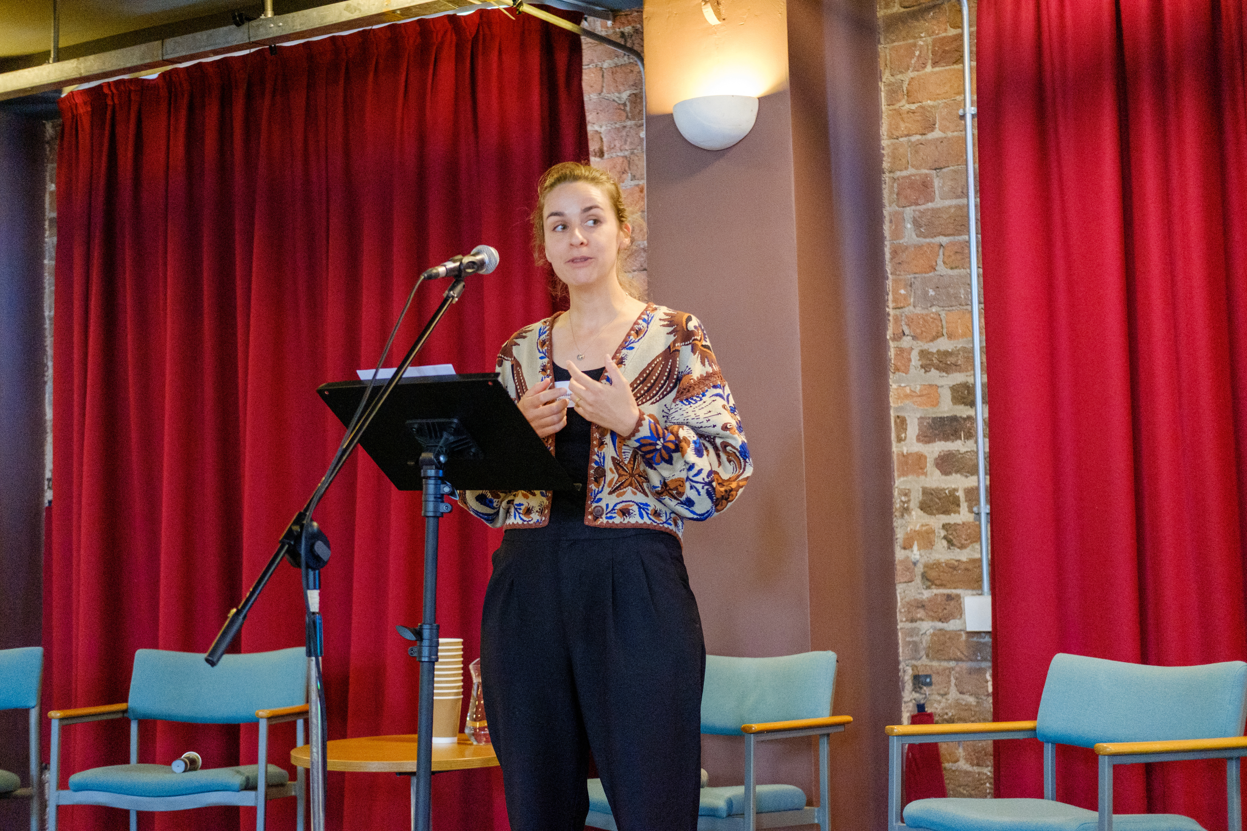 Justine Boussard standing on a stage with large red curtains behind a microphone stand.