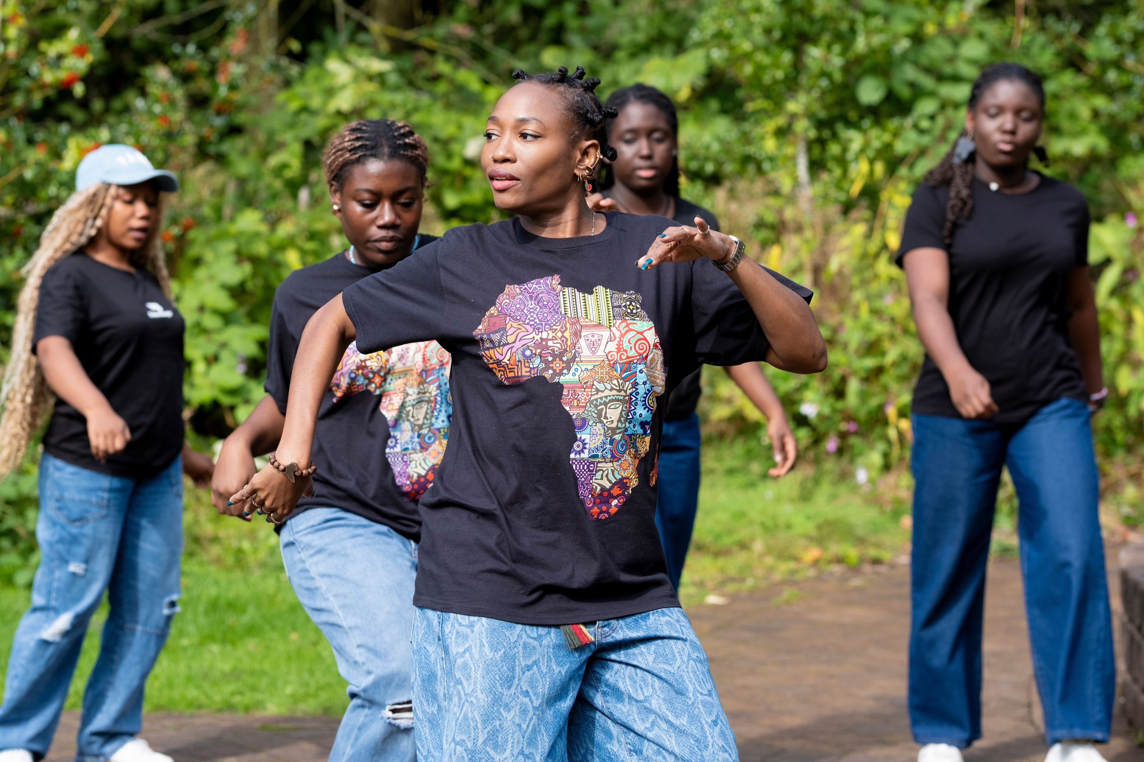 A group of young people dancing. All of them are wearing jeans and a black top with a pattern.