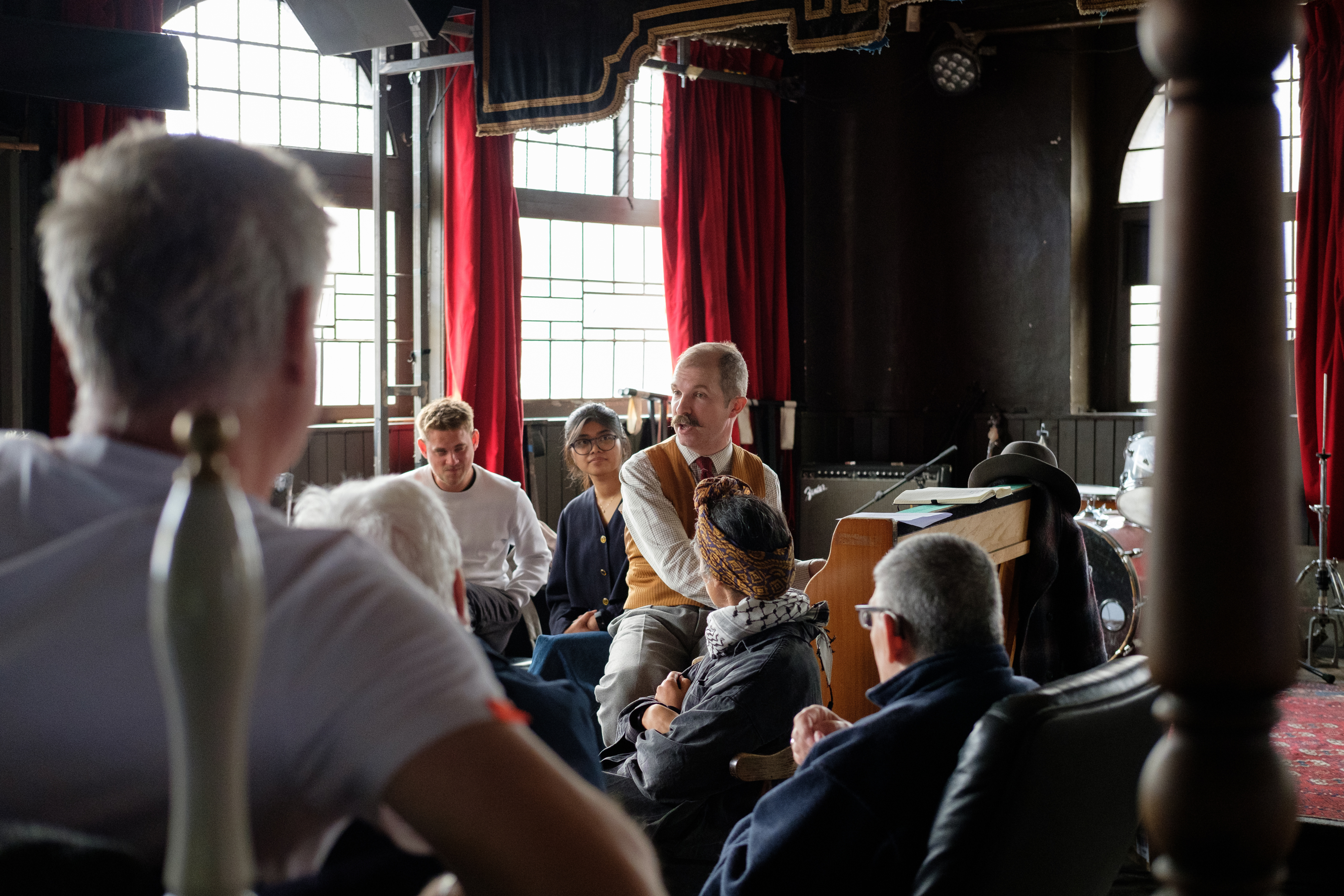 A group of elderly men sitting around in an old-style pub with dark wooden benches and big windows. One person in the middle is playing the piano.