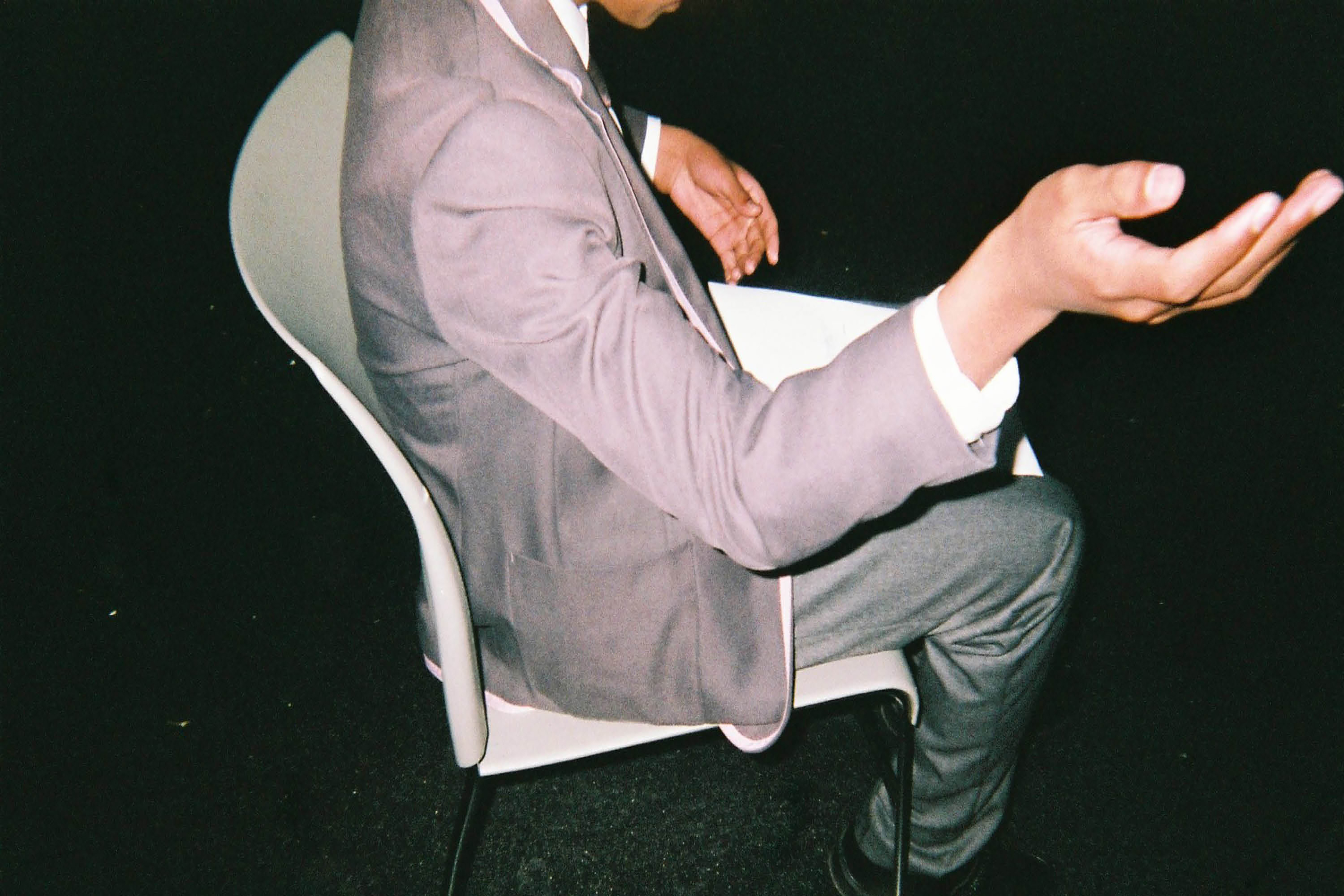 A photograph of a young male wearing school uniform sitting on a chair in a dark room with his arm raised up.