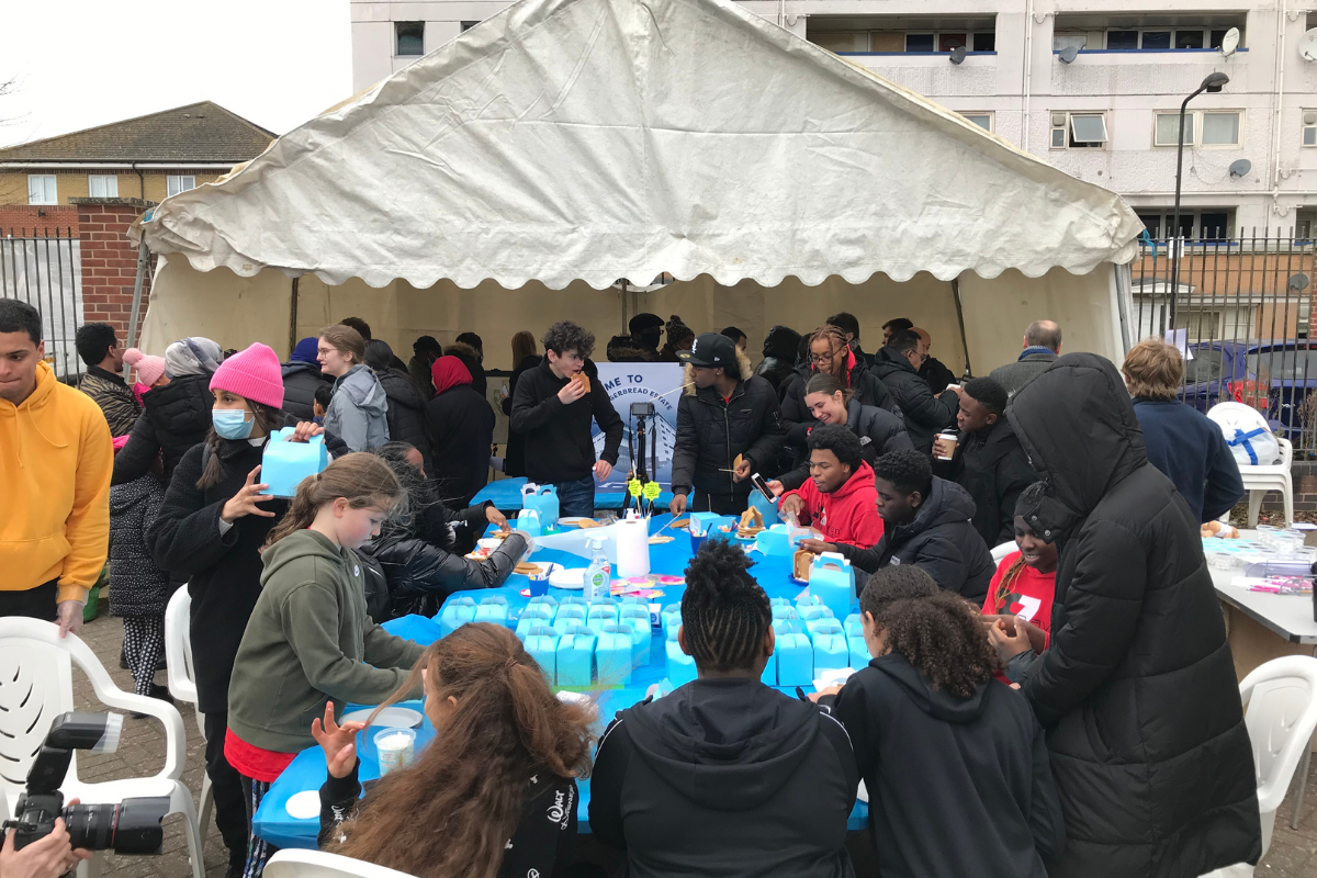 A large table covered in a blue plastic sheet with multiple blue boxes, gingerbread houses, cake decorating items and children sitting around on white plastic chairs talking, in front of a large cream gazebo.