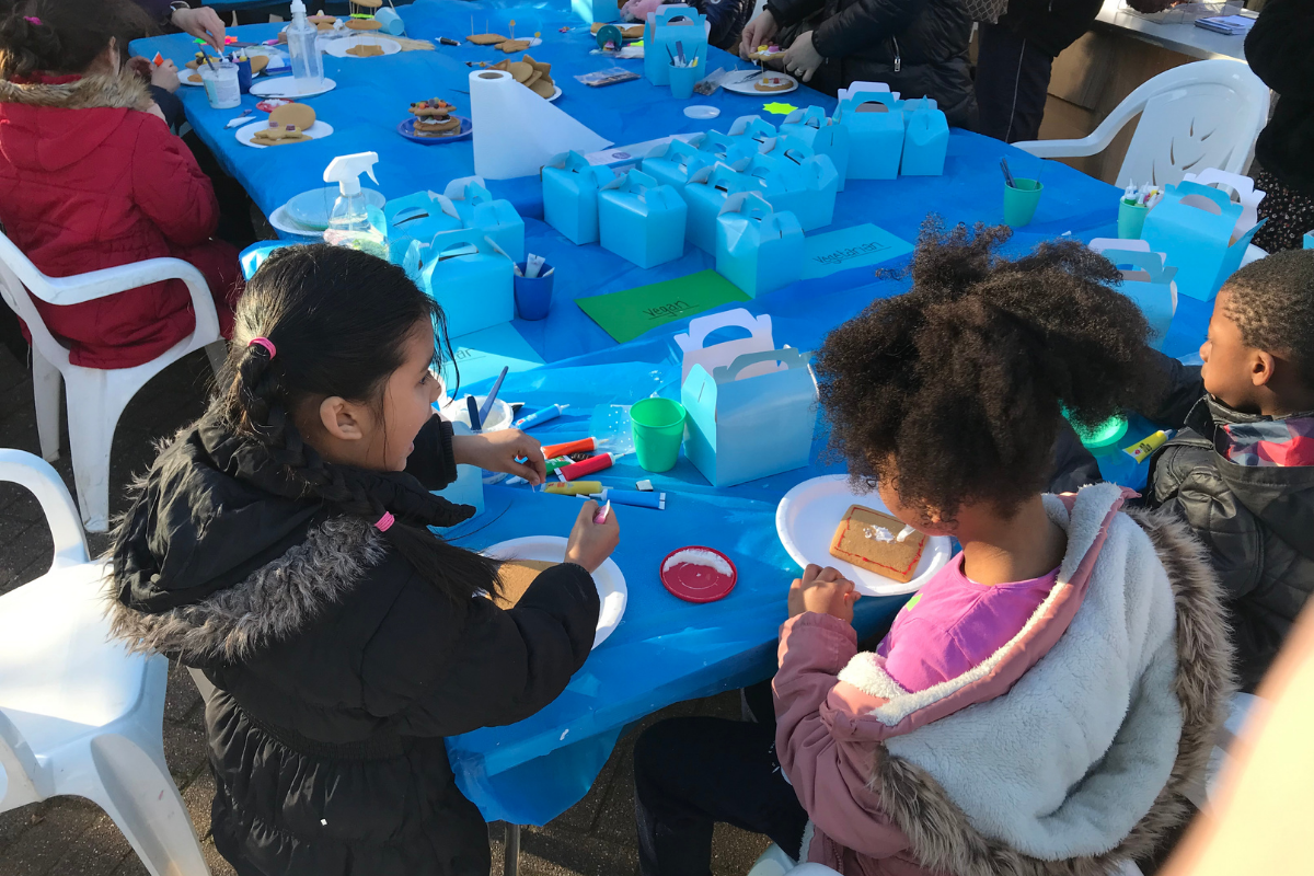 A large table covered in a blue plastic sheet with multiple blue boxes, gingerbread houses, cake decorating items and children sitting around on white plastic chairs talking.