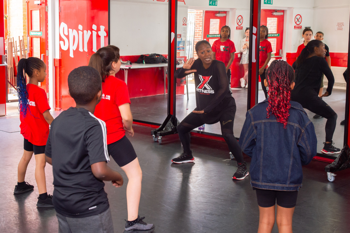 A group of children dancing in front of an adult showing them the moves, all in a large dance studio in front of a wall of large mirrors.
