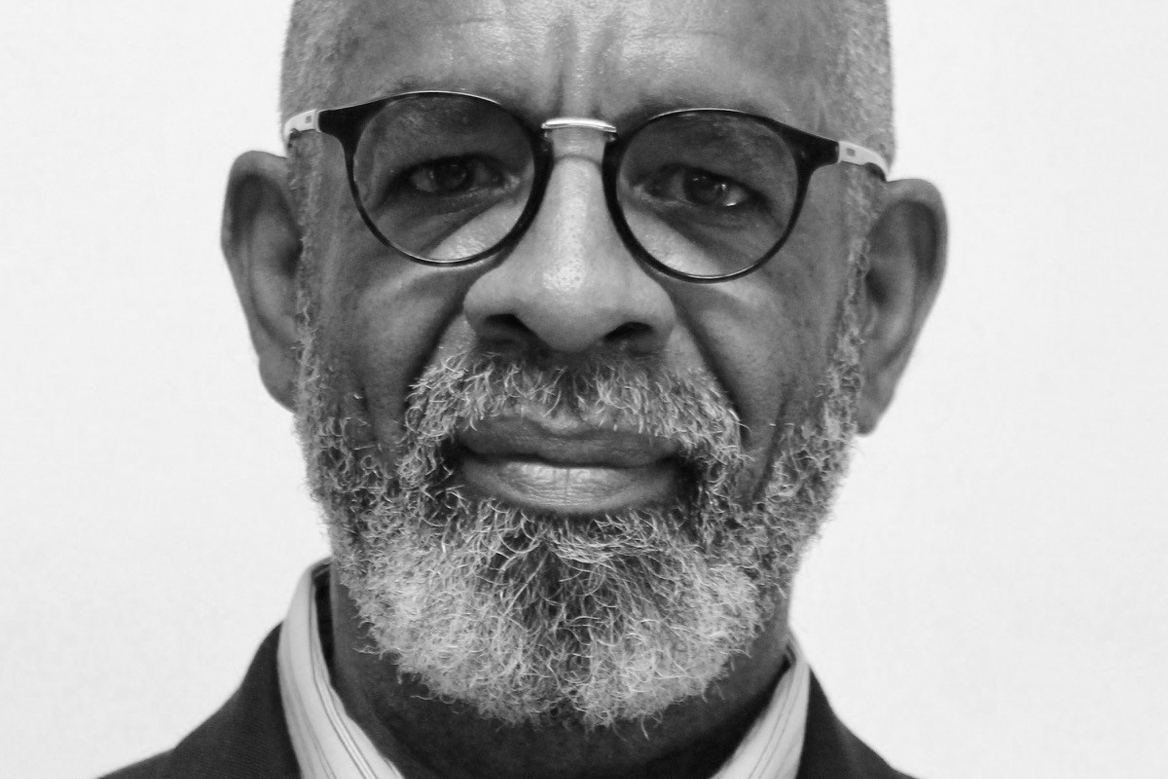 A close-up black and white photograph of a black man wearing glasses standing in front of a plain white background.
