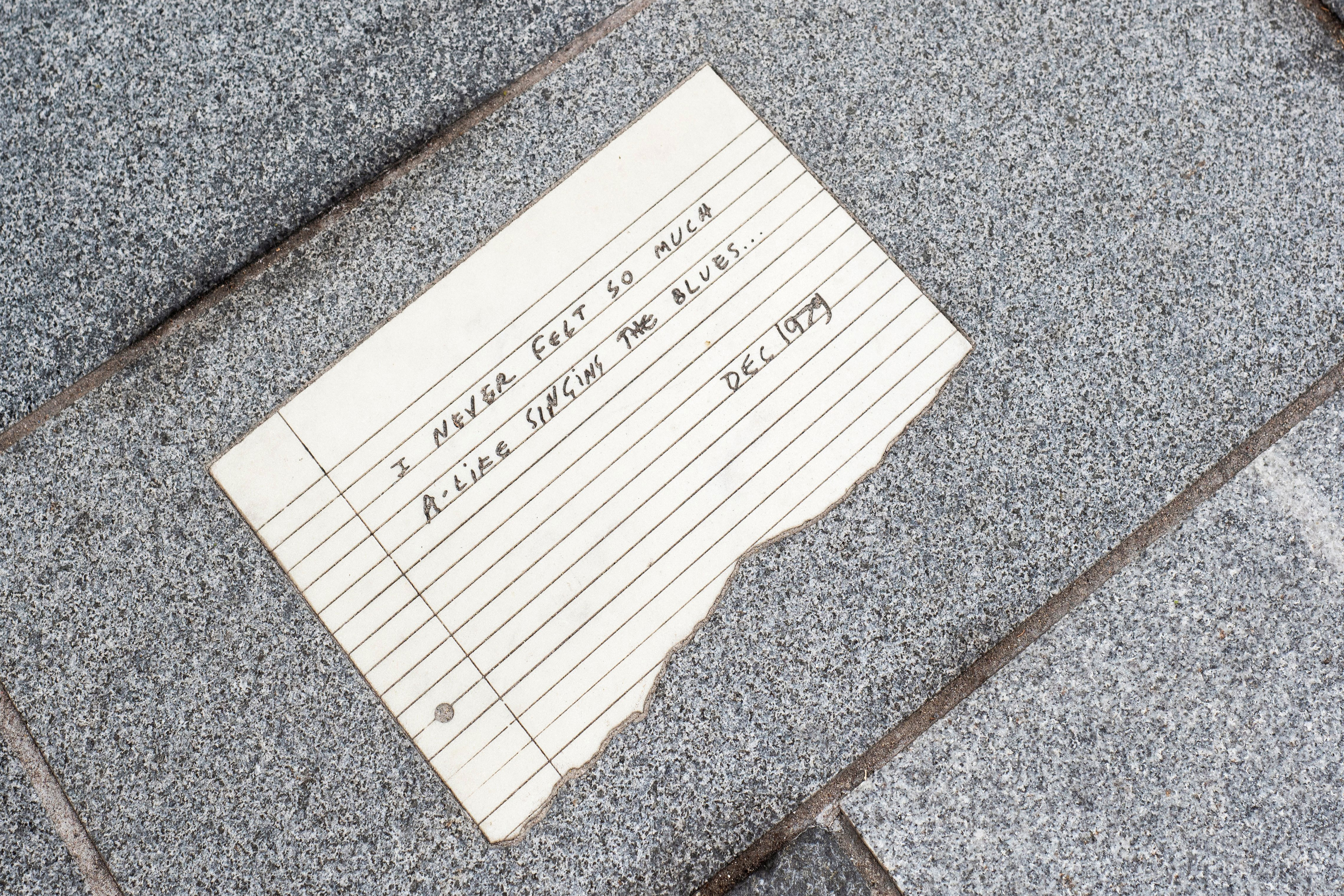 A photograph of a limestone floor segment engraved to replicate an A4 lined paper with a message that reads "I never felt so much A-life singing the blues" dated Dec 1979.