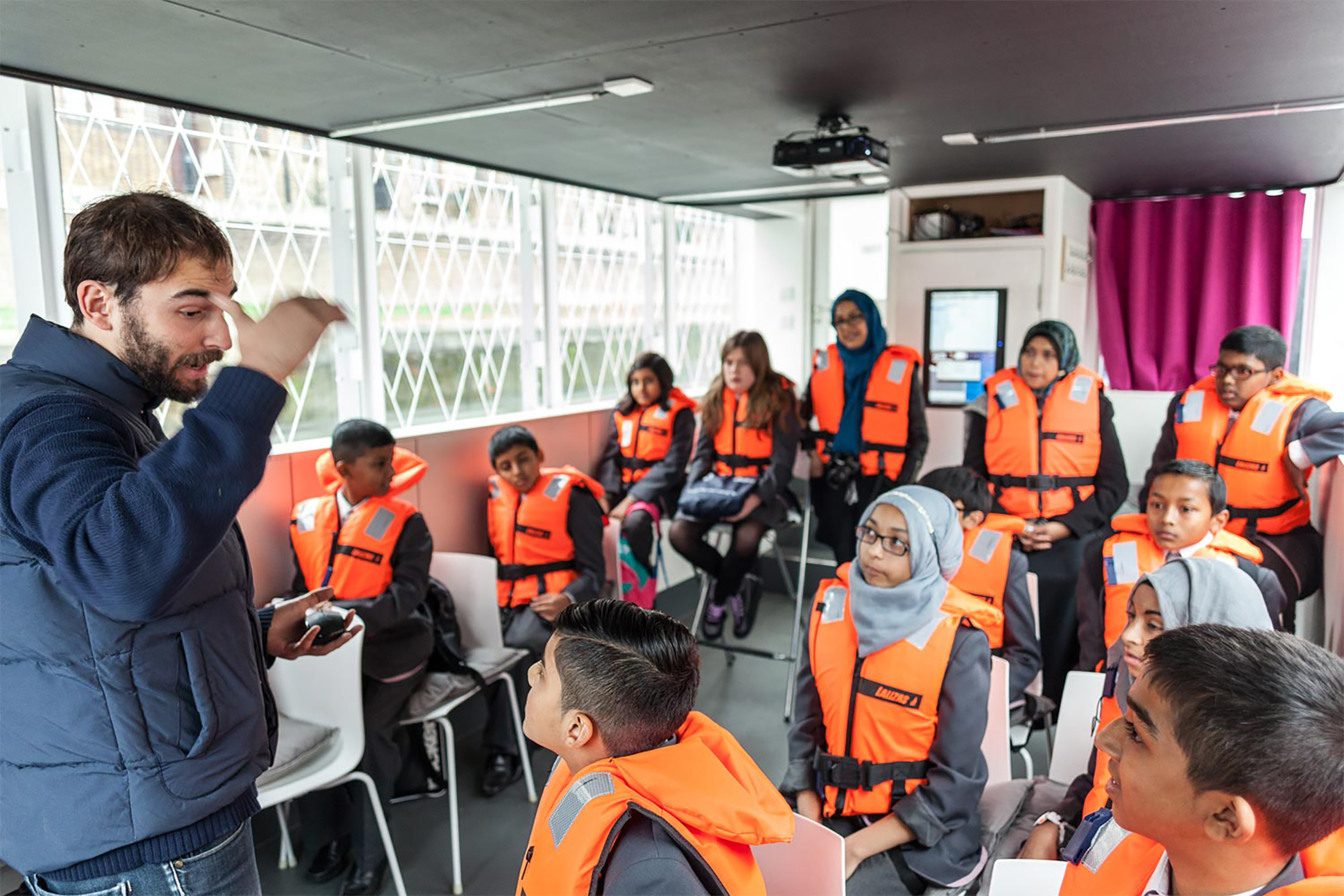 A man wearing a blue jacket teaching a class full of school children wearing orange life jackets inside the room of boat with large windows.