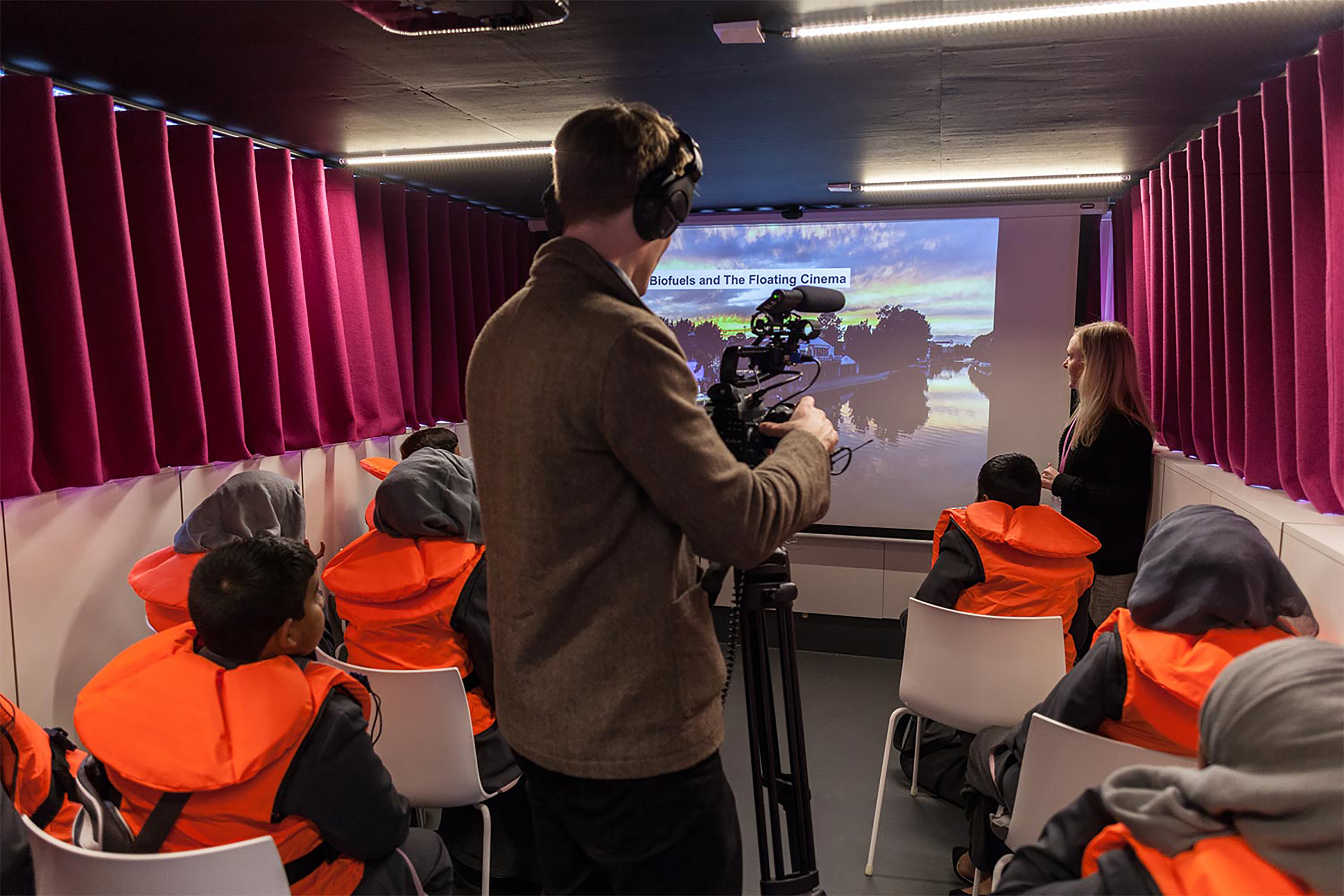 A man wearing a brown jacket and headphones using a camera mounted on a tripod, projecting a film onto a large screen watched by students wearing orange life jackets.