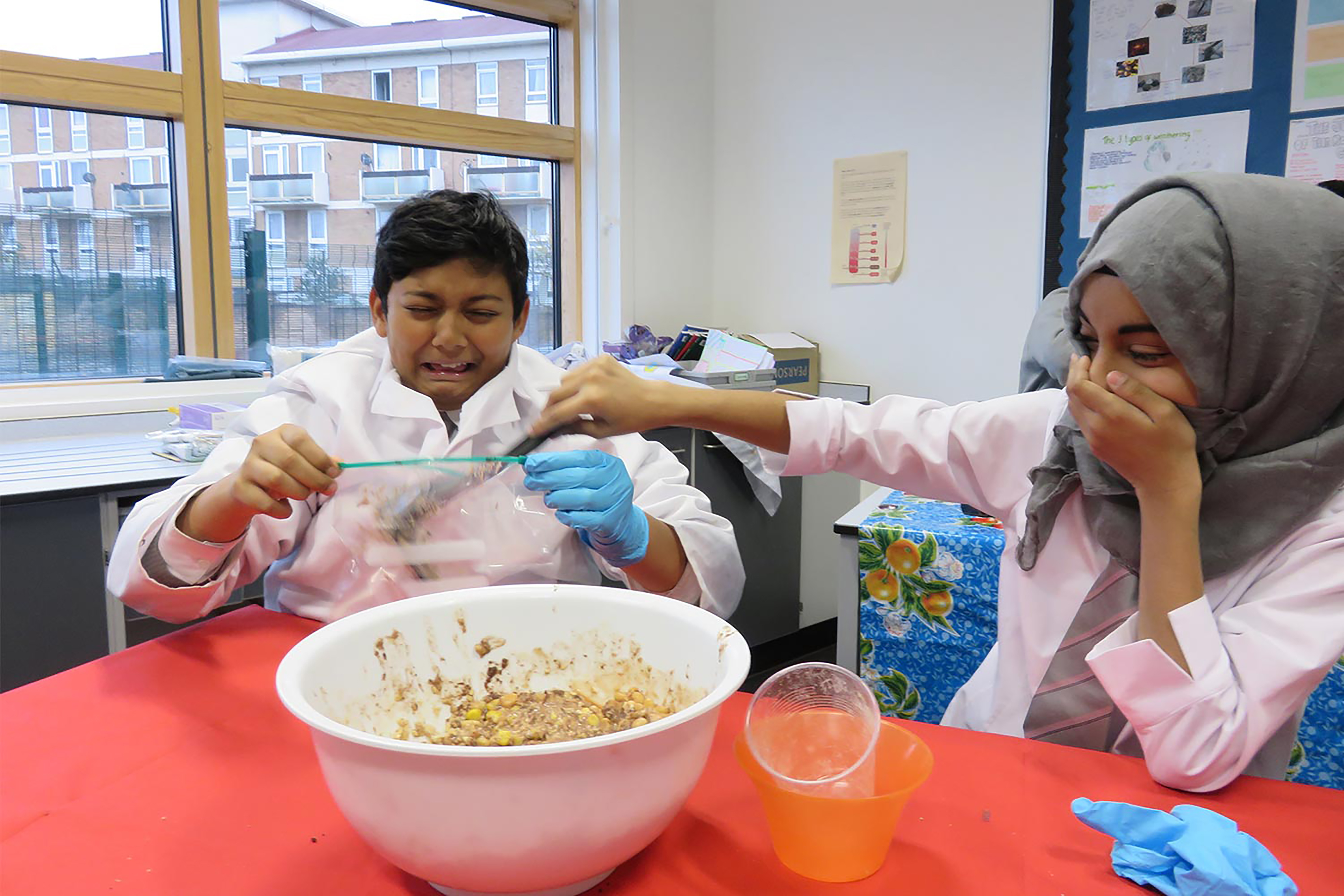 A boy and a girl laughing while making a bowl of paste on a bright red table in a school classroom.