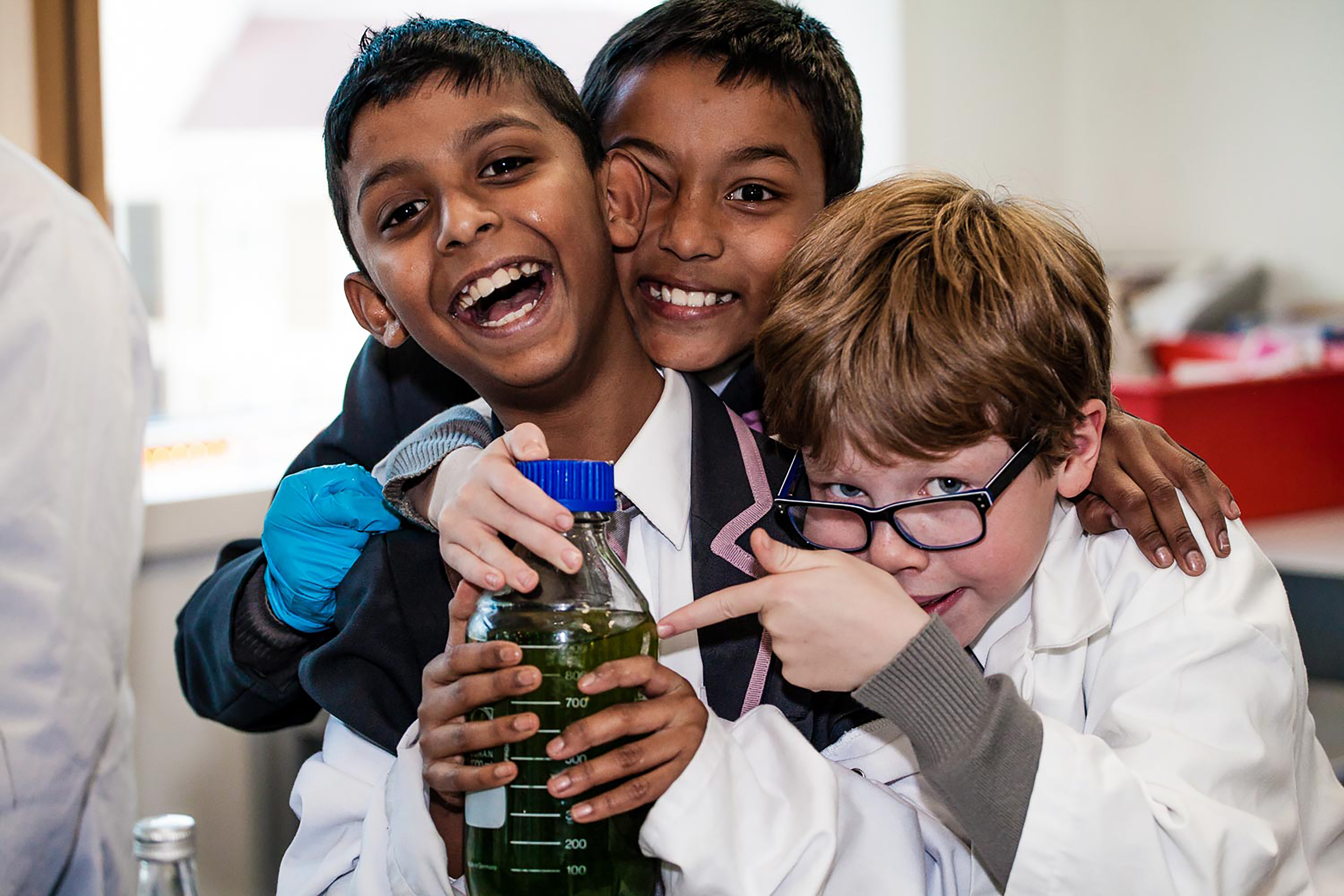 A group of boys laughing and smiling, holding a laboratory bottle and wearing lab coats.
