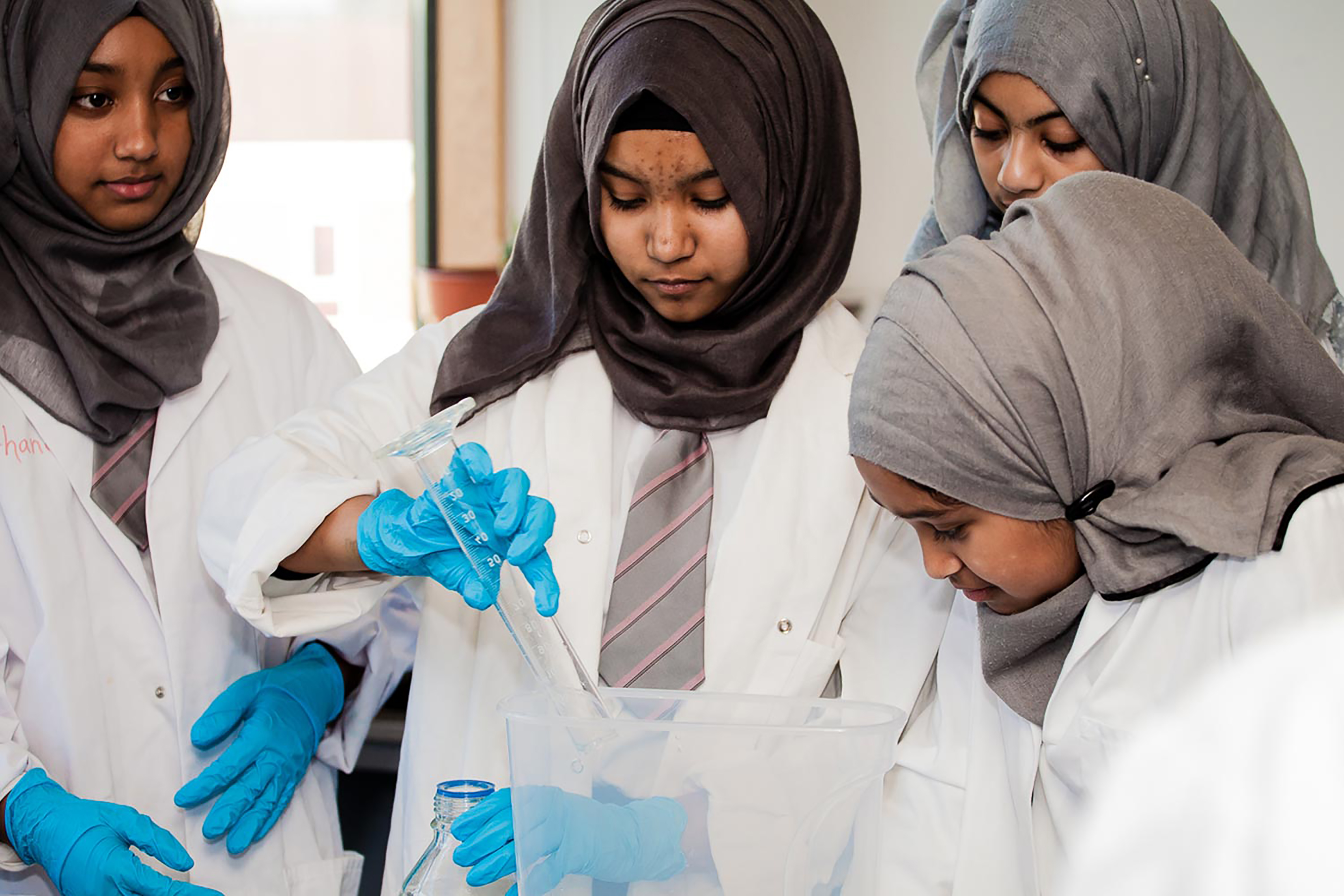 Four girls wearing lab coats and latex gloves pouring liquid into a clear glass mixer.
