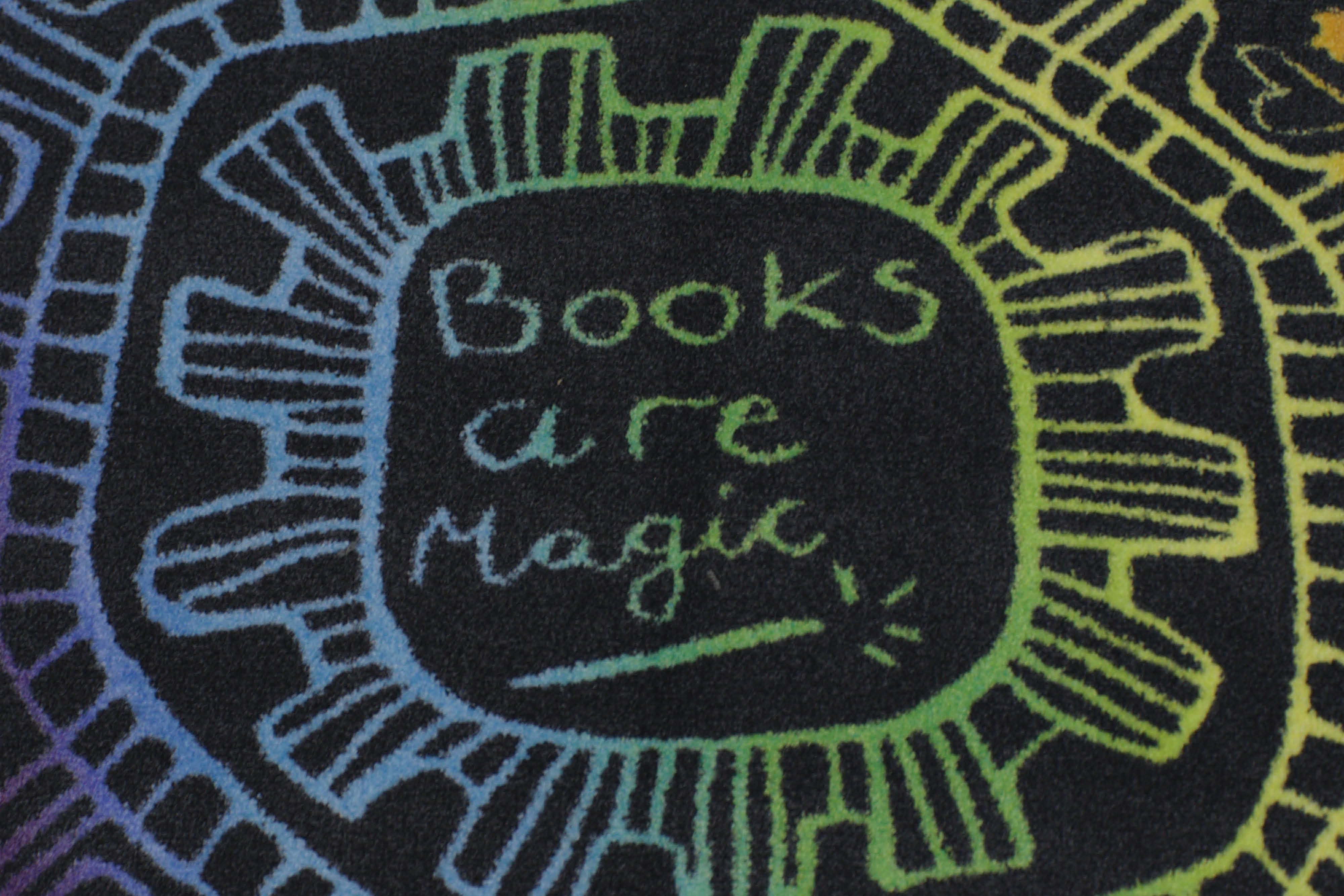 A close up of a section of a rug with a circular repeated pattern in a blue, yellow and green total colour scheme. The circle has the words “Books are magic” in a handwritten style with a drawing of a magic wand.