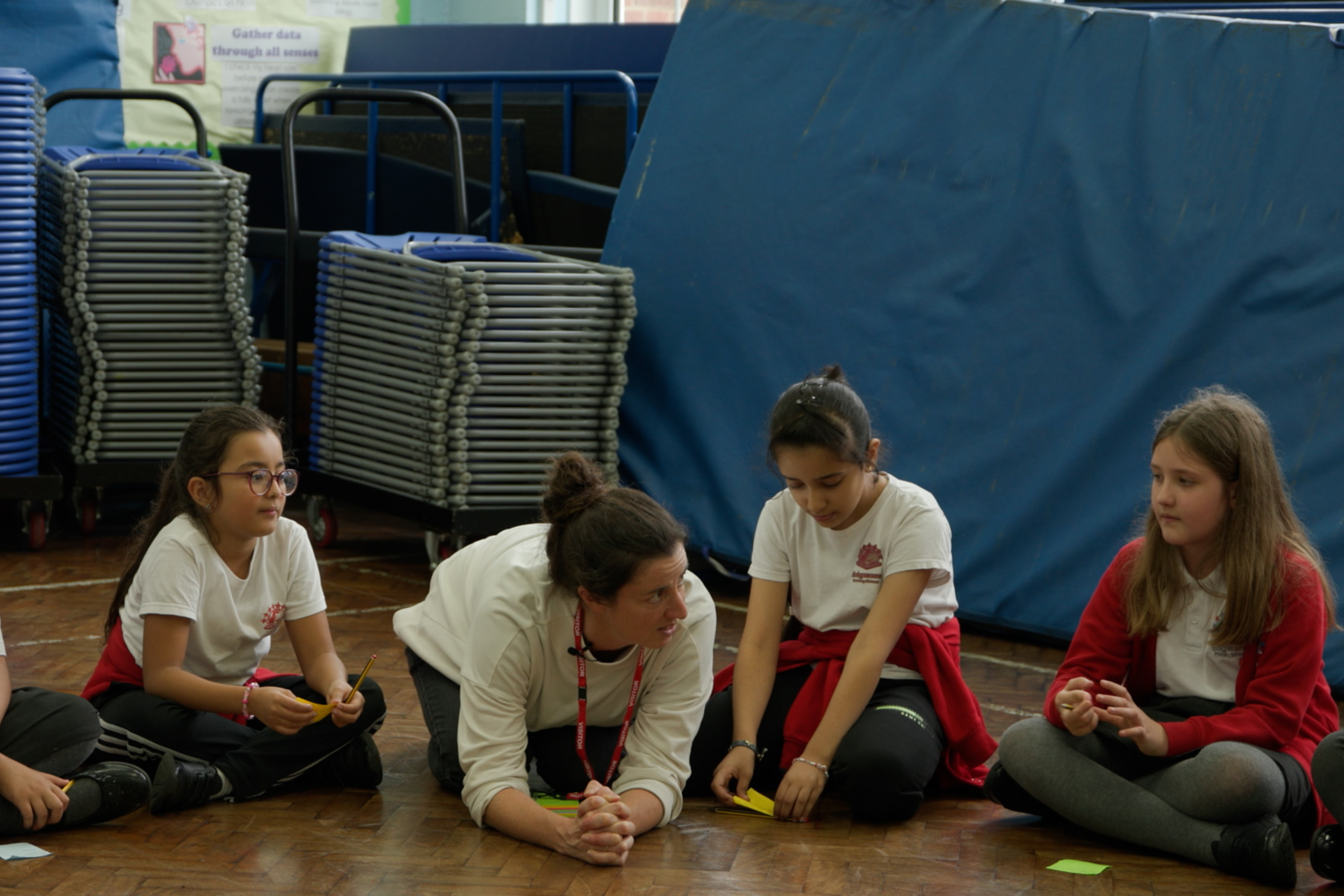 A woman and group of school children sitting in a line talking. They are in a large classroom with objects on the walls.