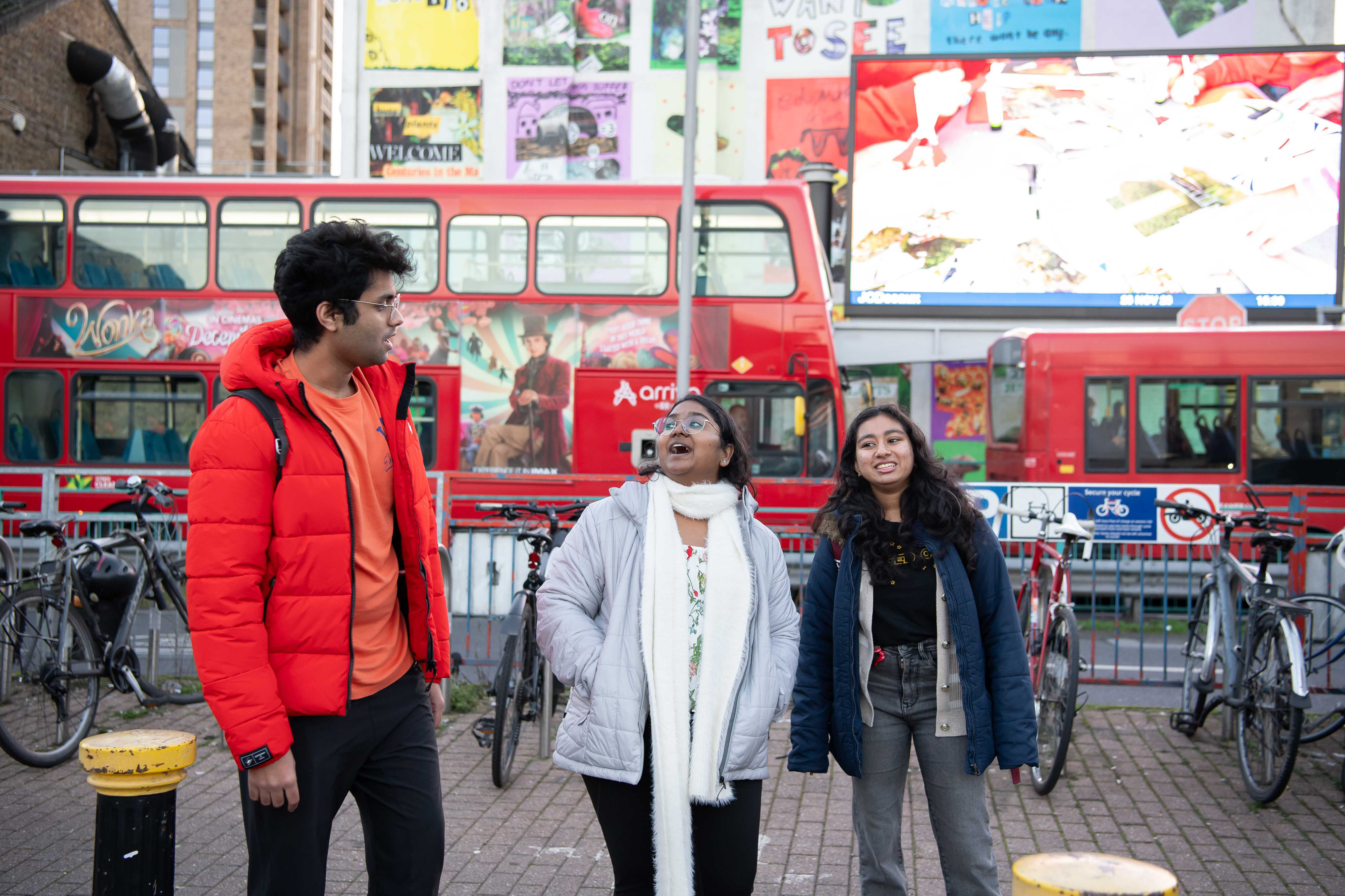 Three people standing on singing. They are all wearing coats and in front of red London buses going past.