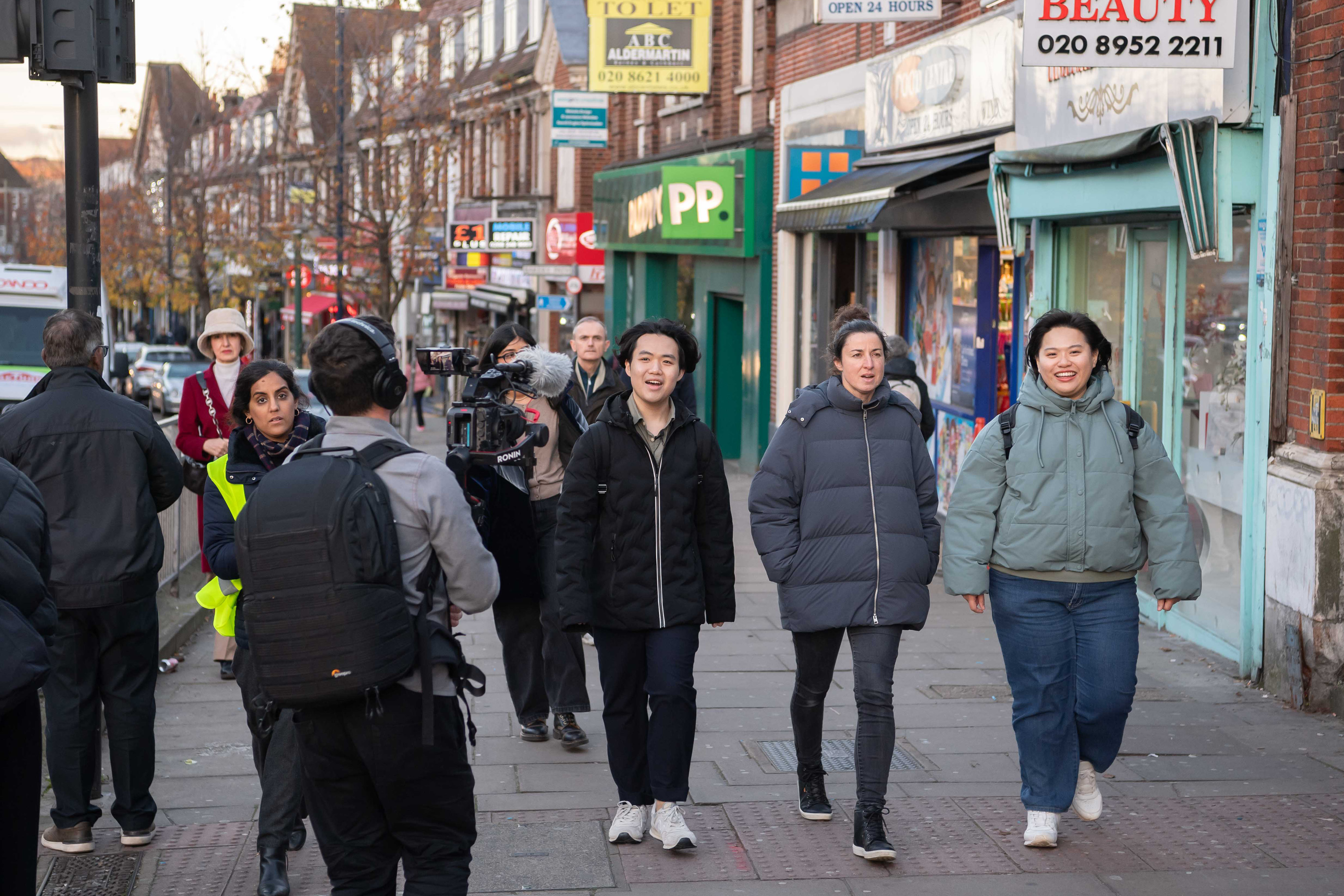 Three people walking down a street singing. They are all wearing coats while a person in front films them.