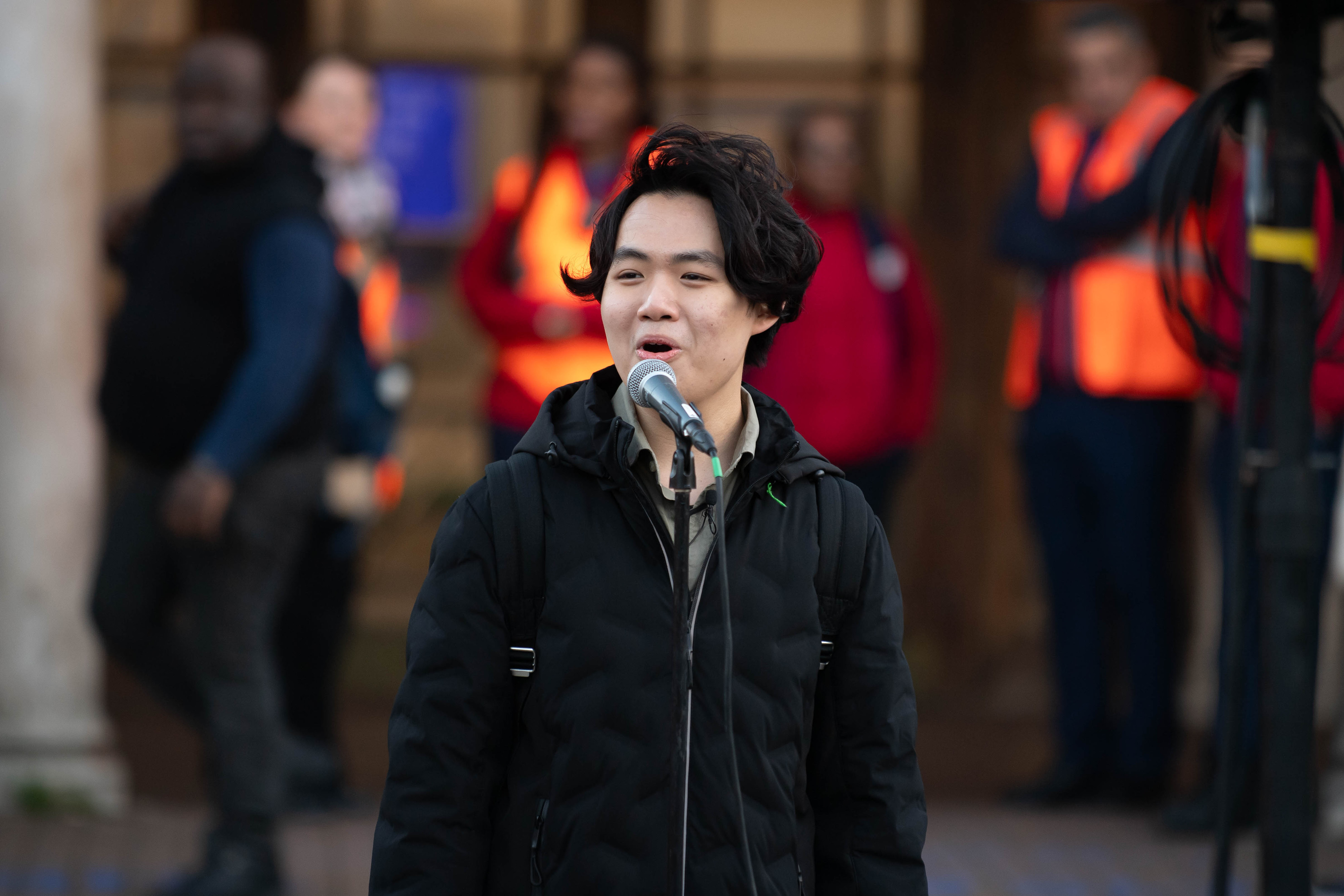 A man singing next to a microphone stand wearing a black top and backpack. He is standing in front of a group of people.