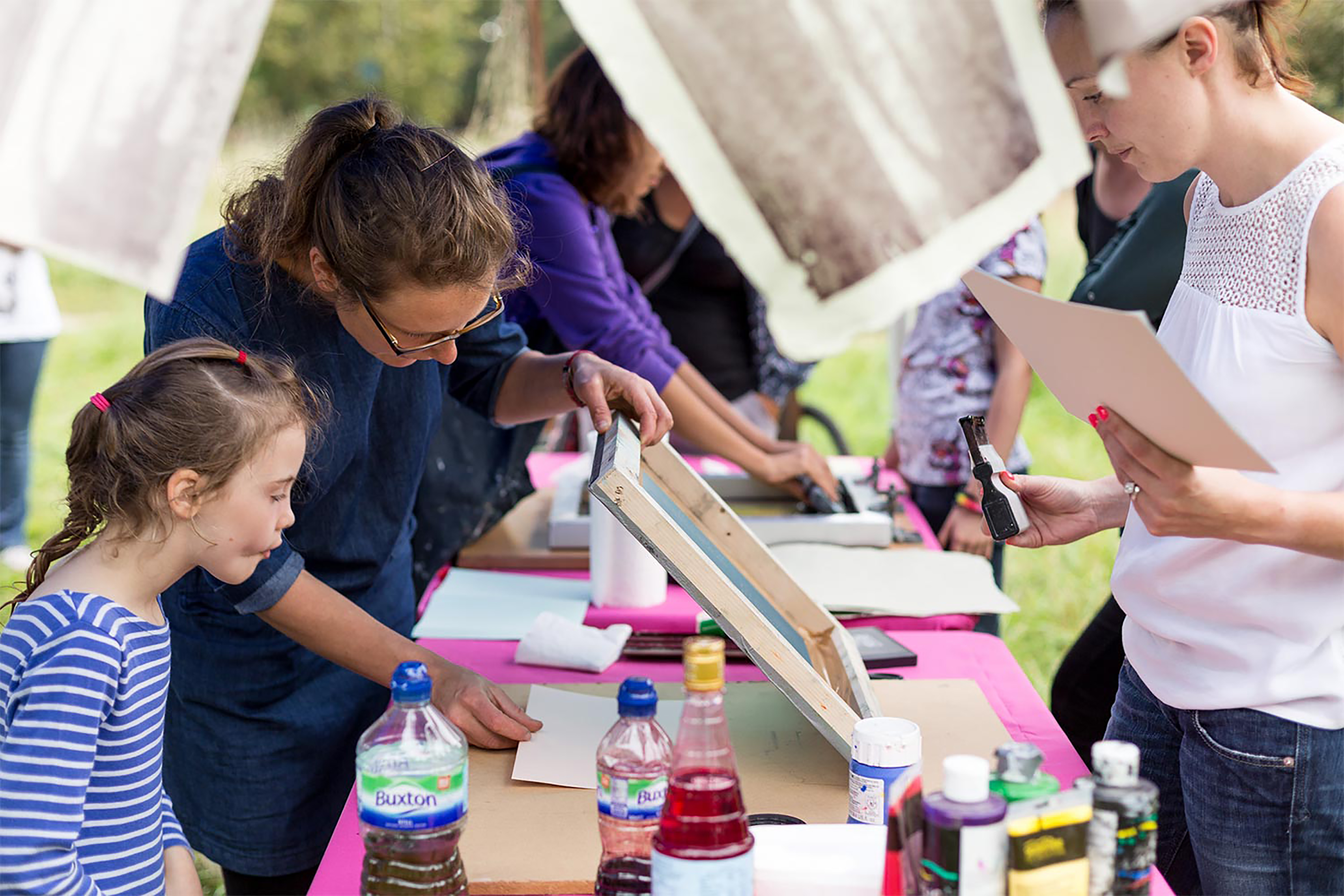 A woman showing a young girl how to screen print machine with images exposed onto it, and a large washing line with prints handing from it.