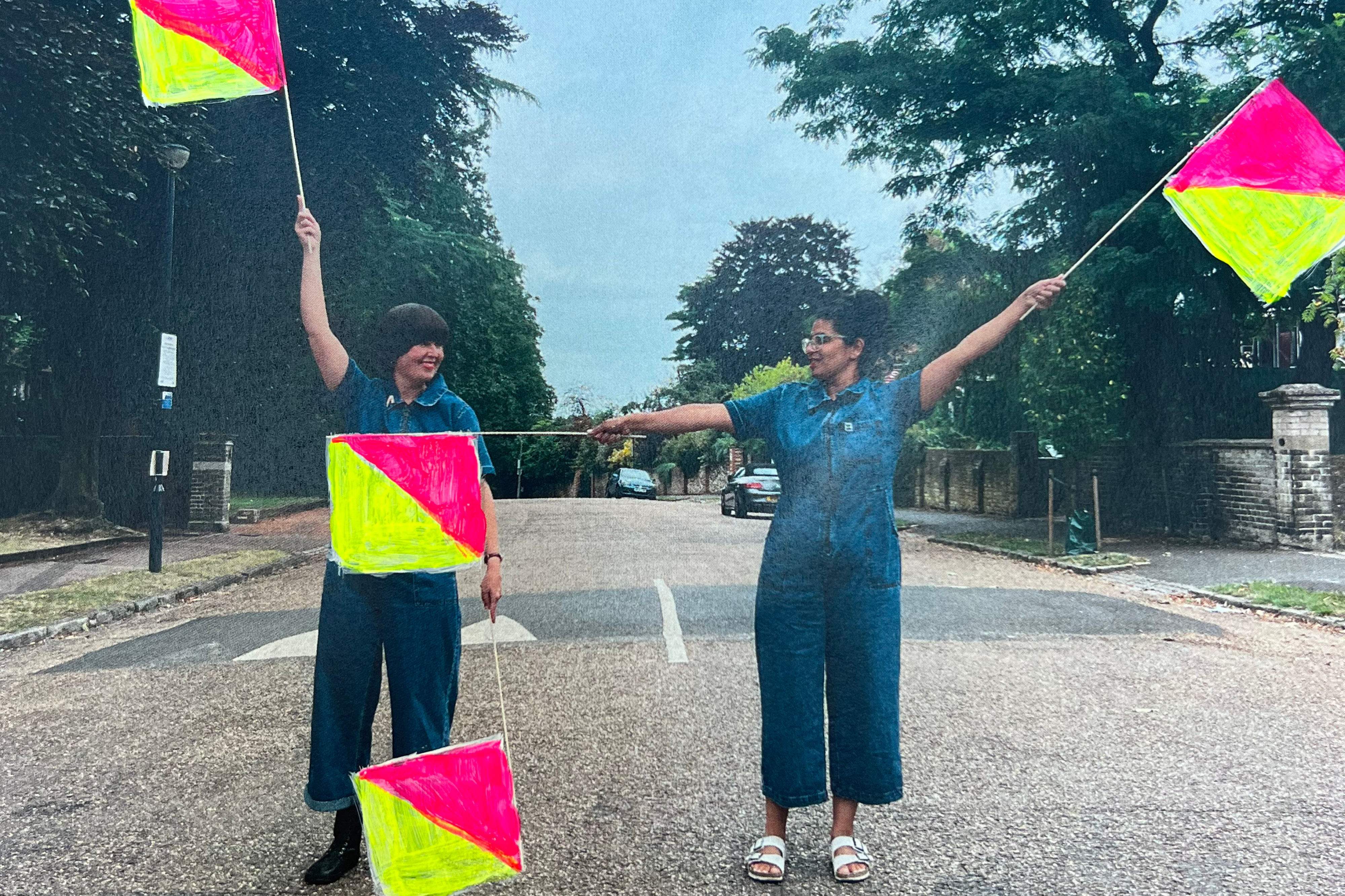 Two people wearing blue jumpsuits standing in a road. They are both holding up two yellow and red triangle flags in the air.
