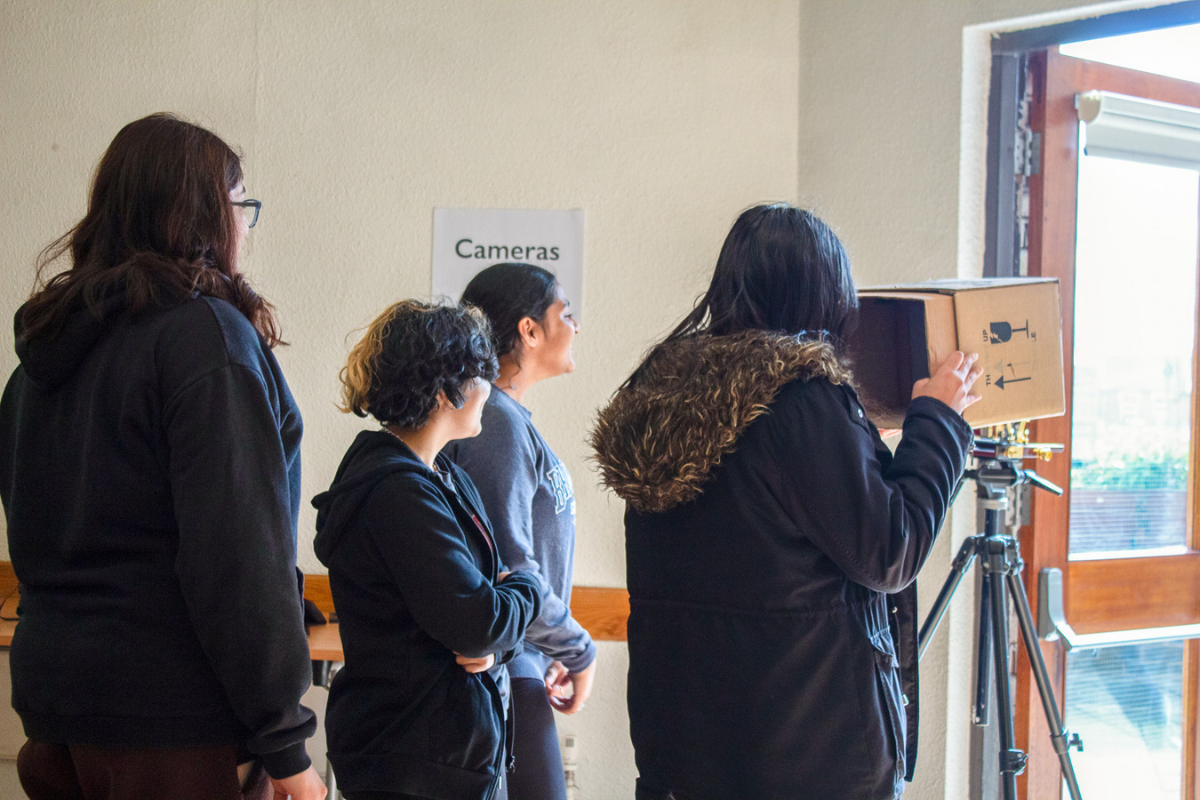 A group a woman looking outside of a door, with one woman holding onto a large rectangular camera on a tripod.