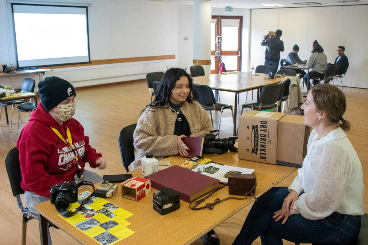 Three people sitting round a large wooden table full of different items, in a blank room with people in the background.