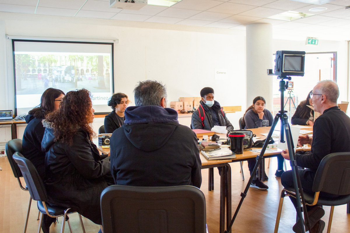 A group of people sitting round a large wooden table full of different items, in a blank room with a large projector screen in the background.