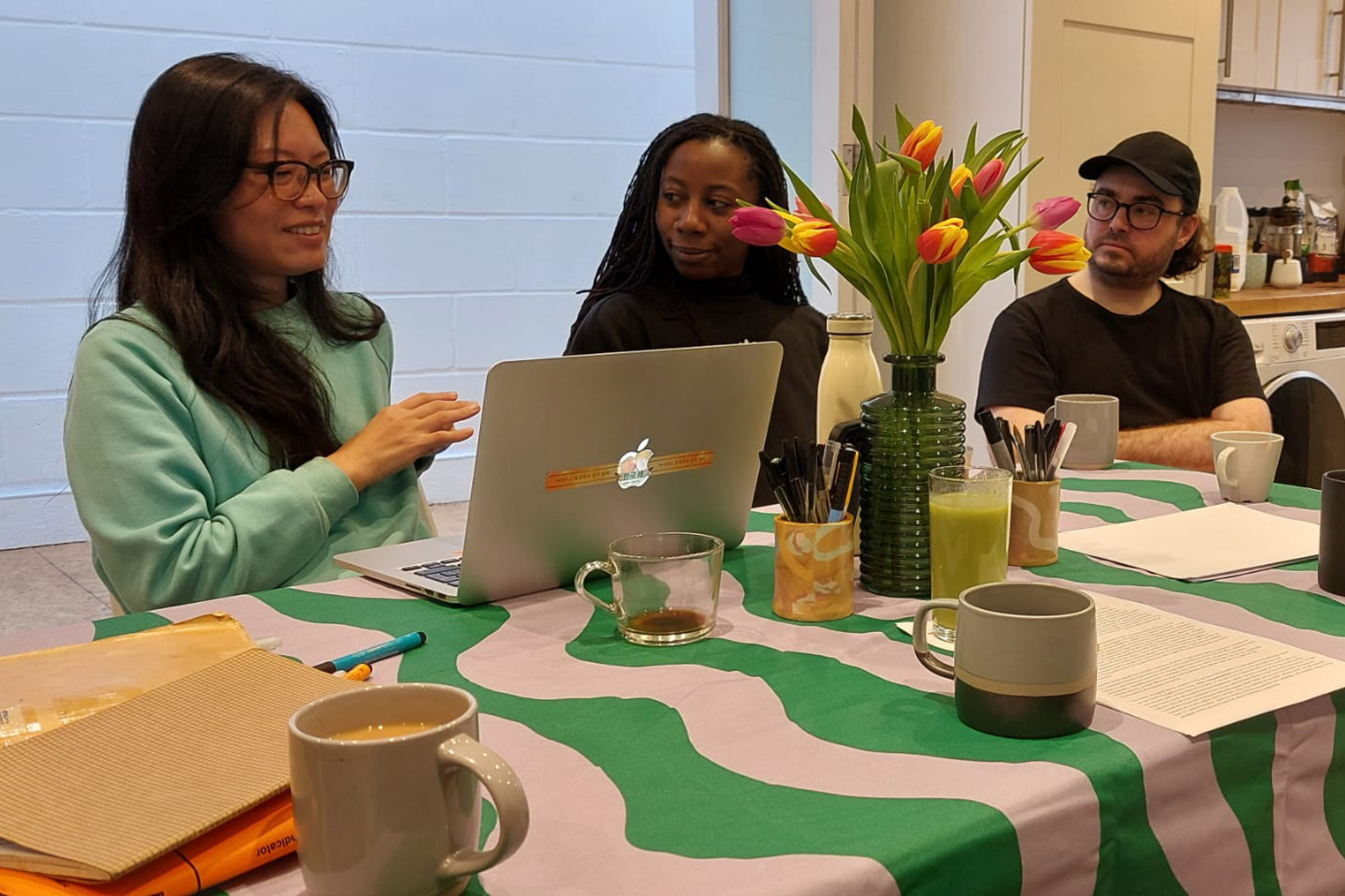 Three people sitting around a table which is decorated with a green and pink patterned tablecloth, various coffee mugs, flowers and notebooks.