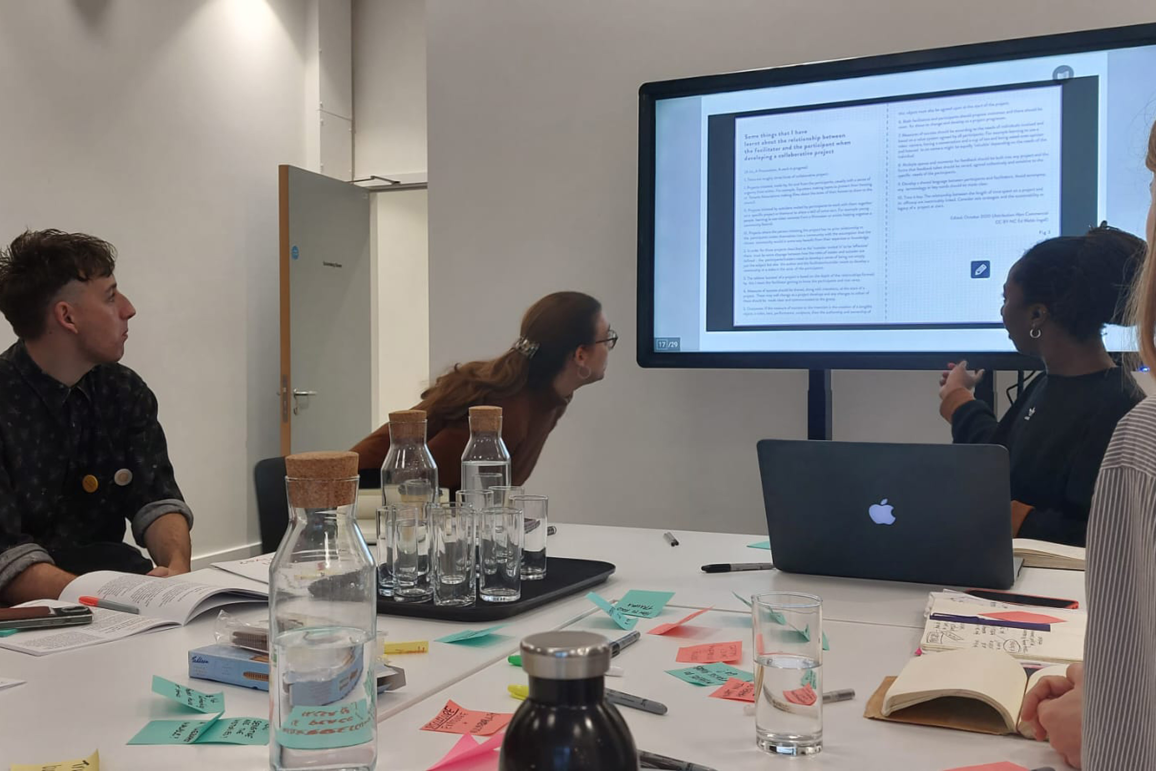 People sitting around a table with colourful sticky notes, water glasses and jugs, laptops and notebooks. They are all facing a large presentation screen with a book being shown.