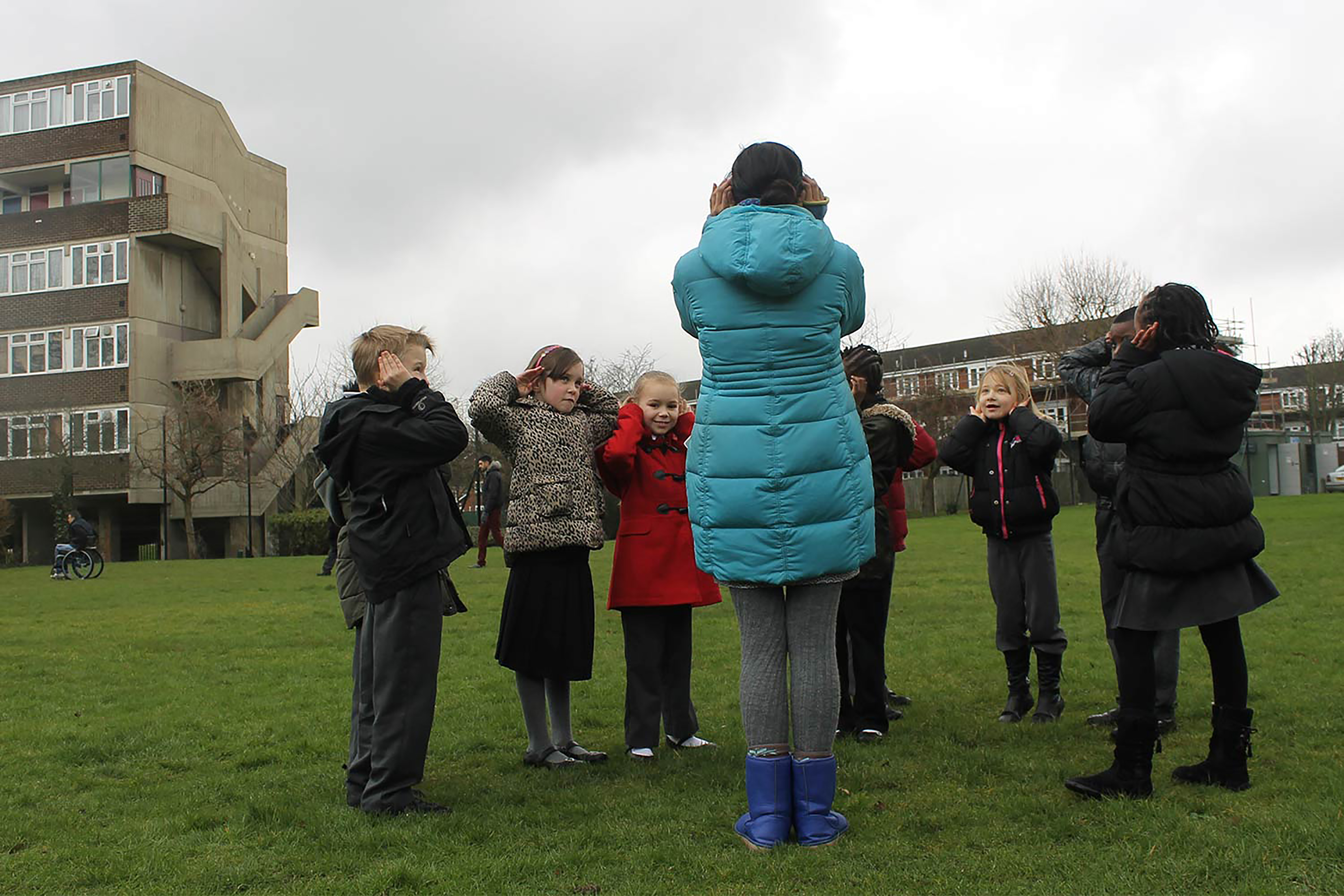 A group of school children in a semi-circle around an adult with their hands over their ears.