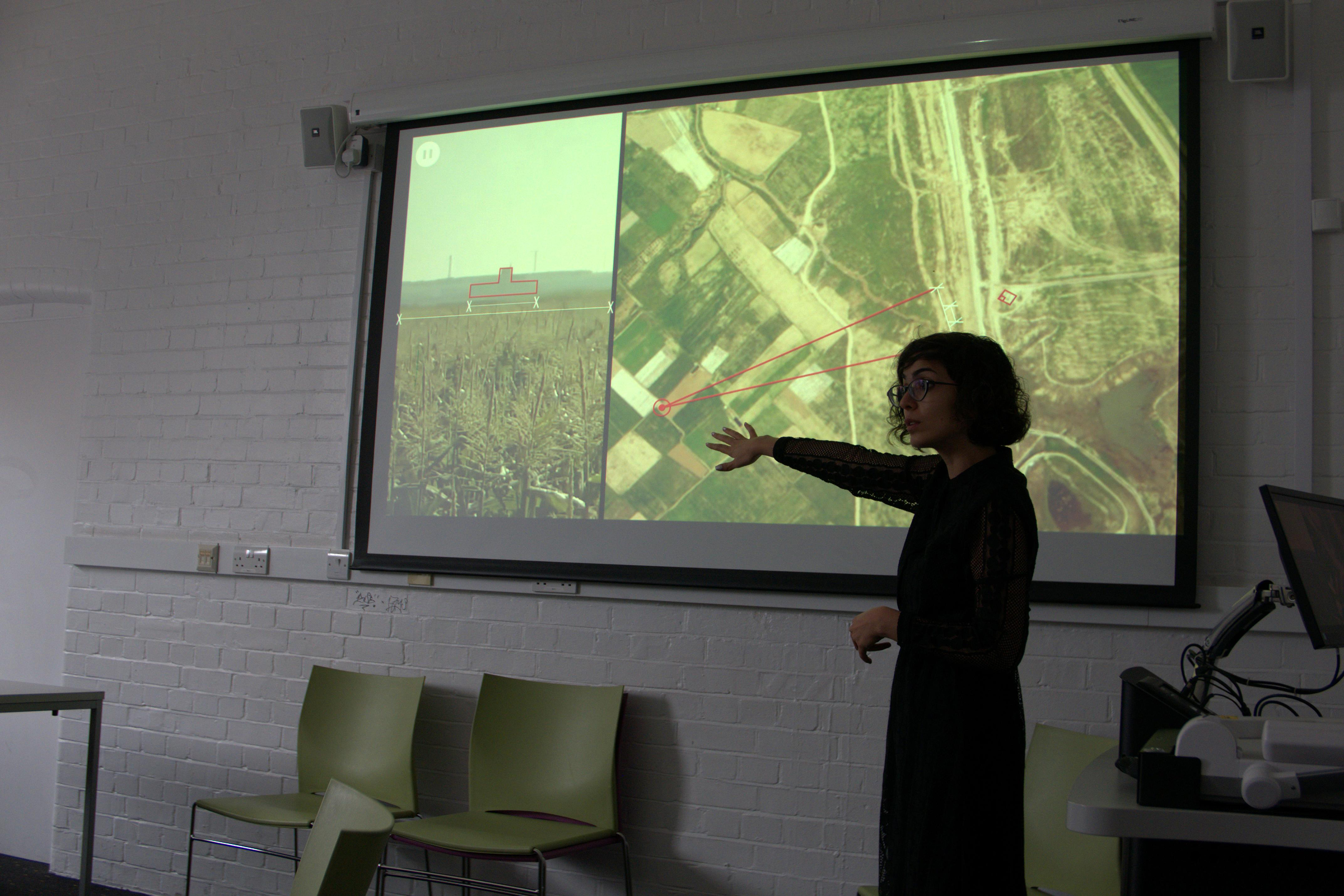 A woman pointing to a bird- eye view of a field projected onto a presentation slide in a classroom.