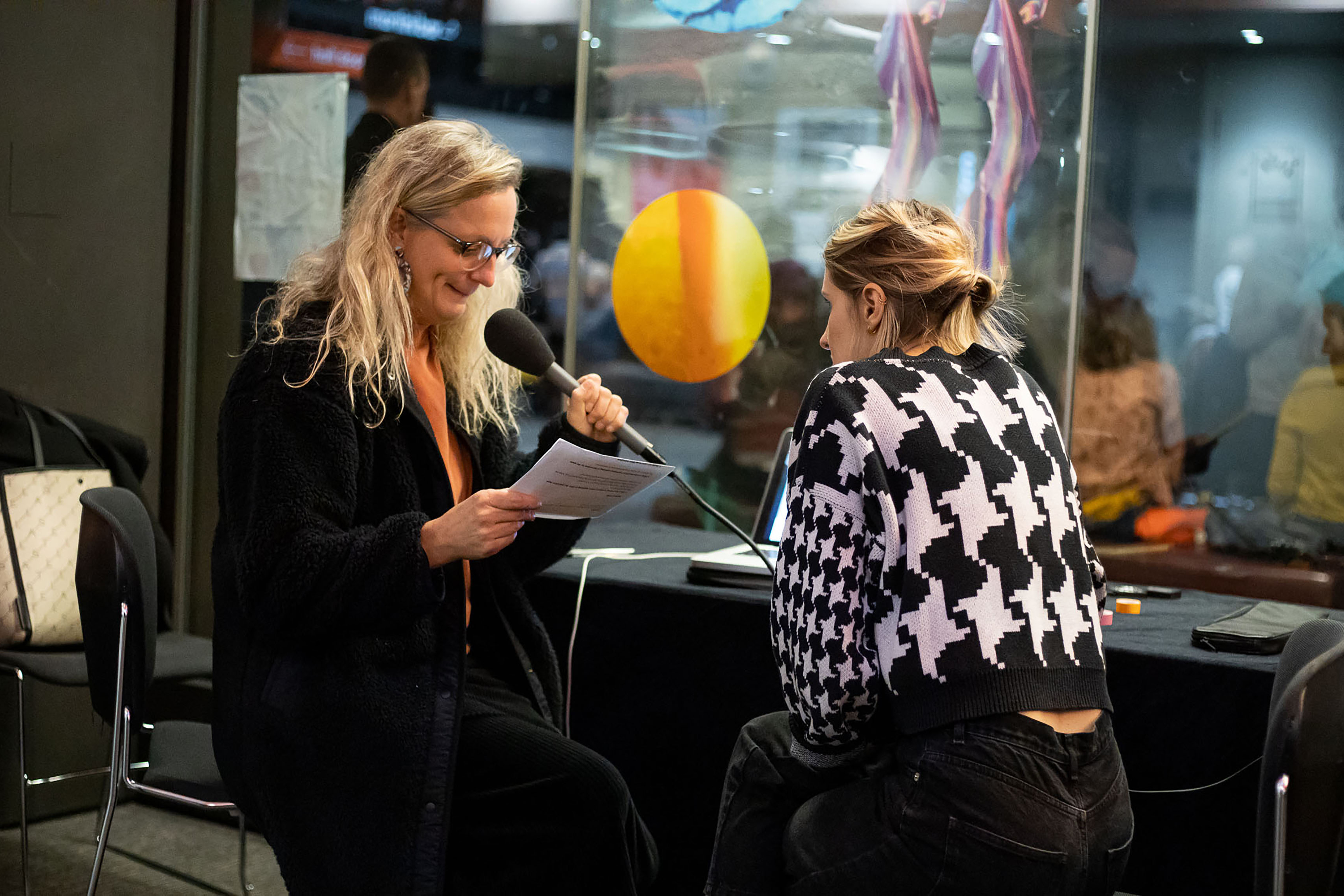 A middle-aged woman holding a microphone while reading from a sheet of paper while another woman watches her in a studio.