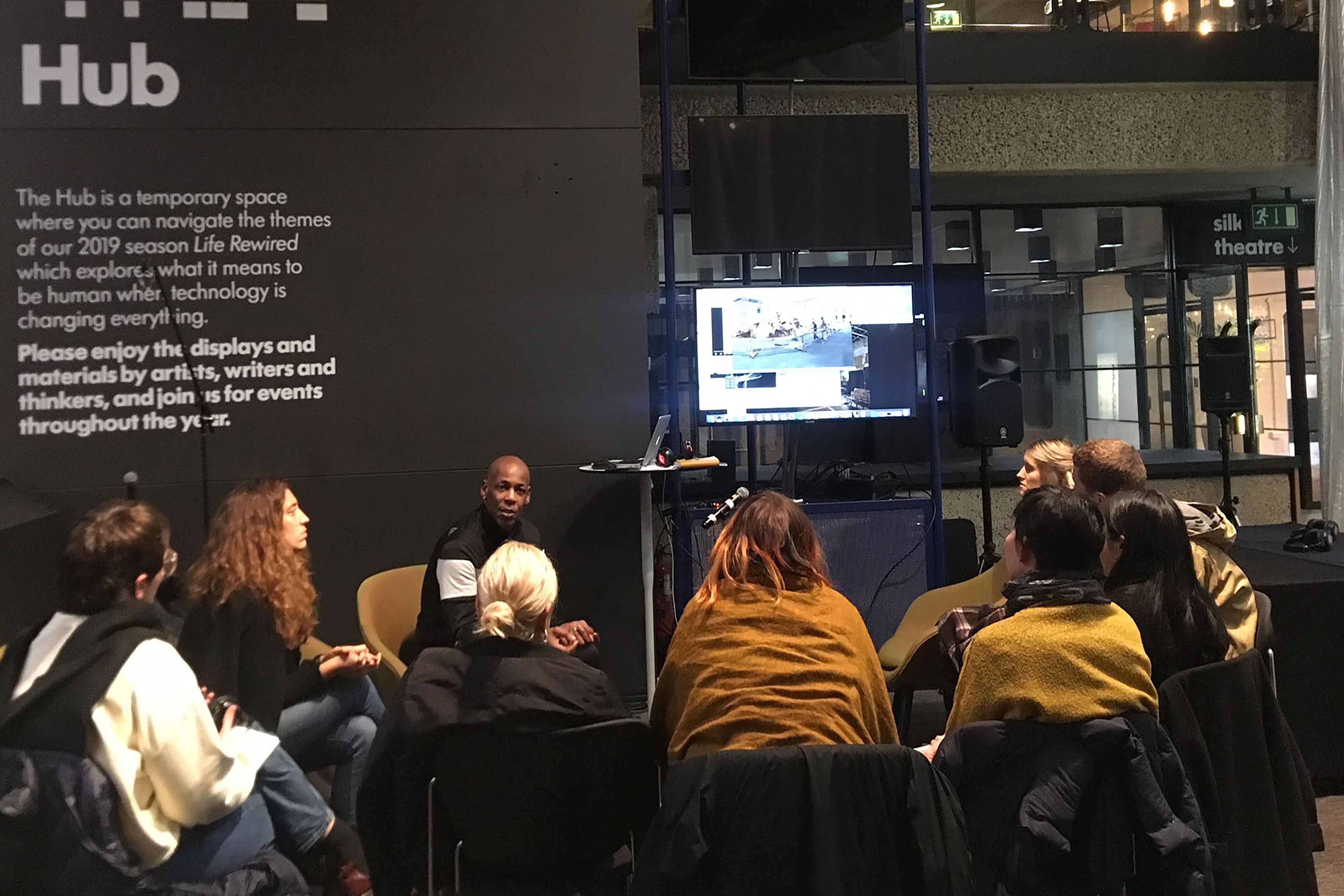 A photograph of a group of people sitting on chairs forming a semi-circle to watch a TV in a dark gallery.