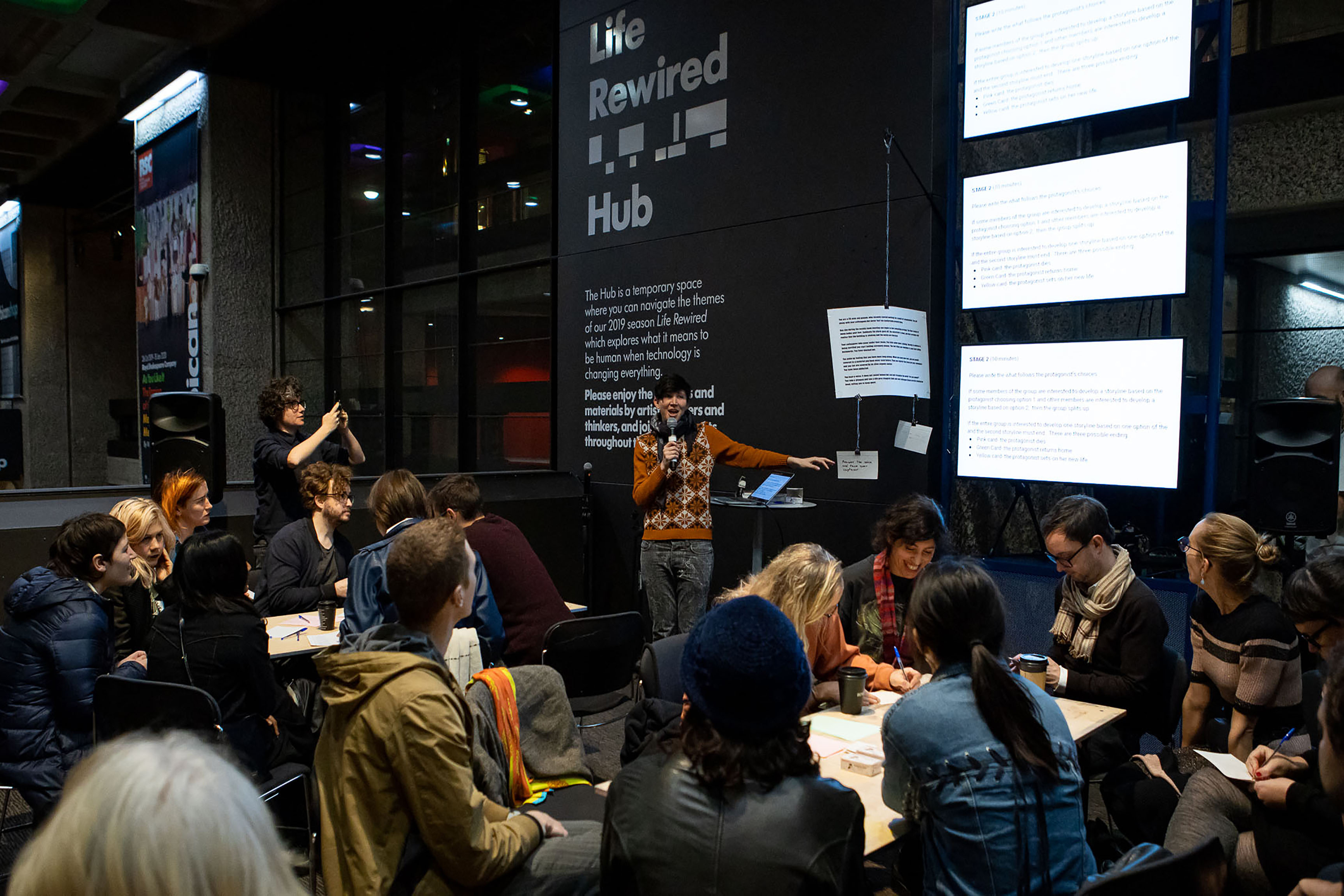 A photograph of a person wearing a knitted sweatshirt holding a microphone and addressing a crowd sat at tables.
