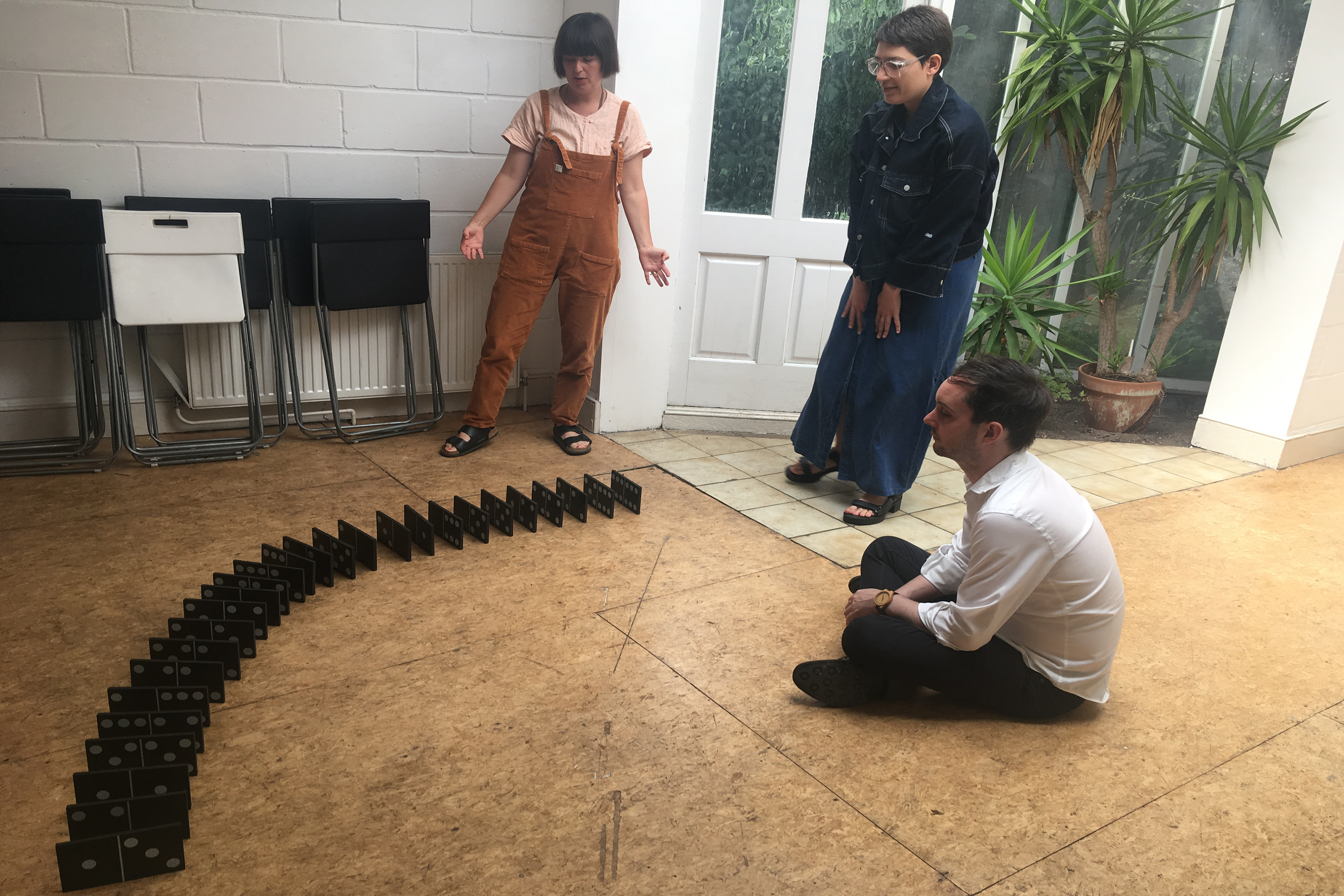 A photograph of a man sat on the floor and two women standing up as they watch a sequence of large dominos fall in a flat house.