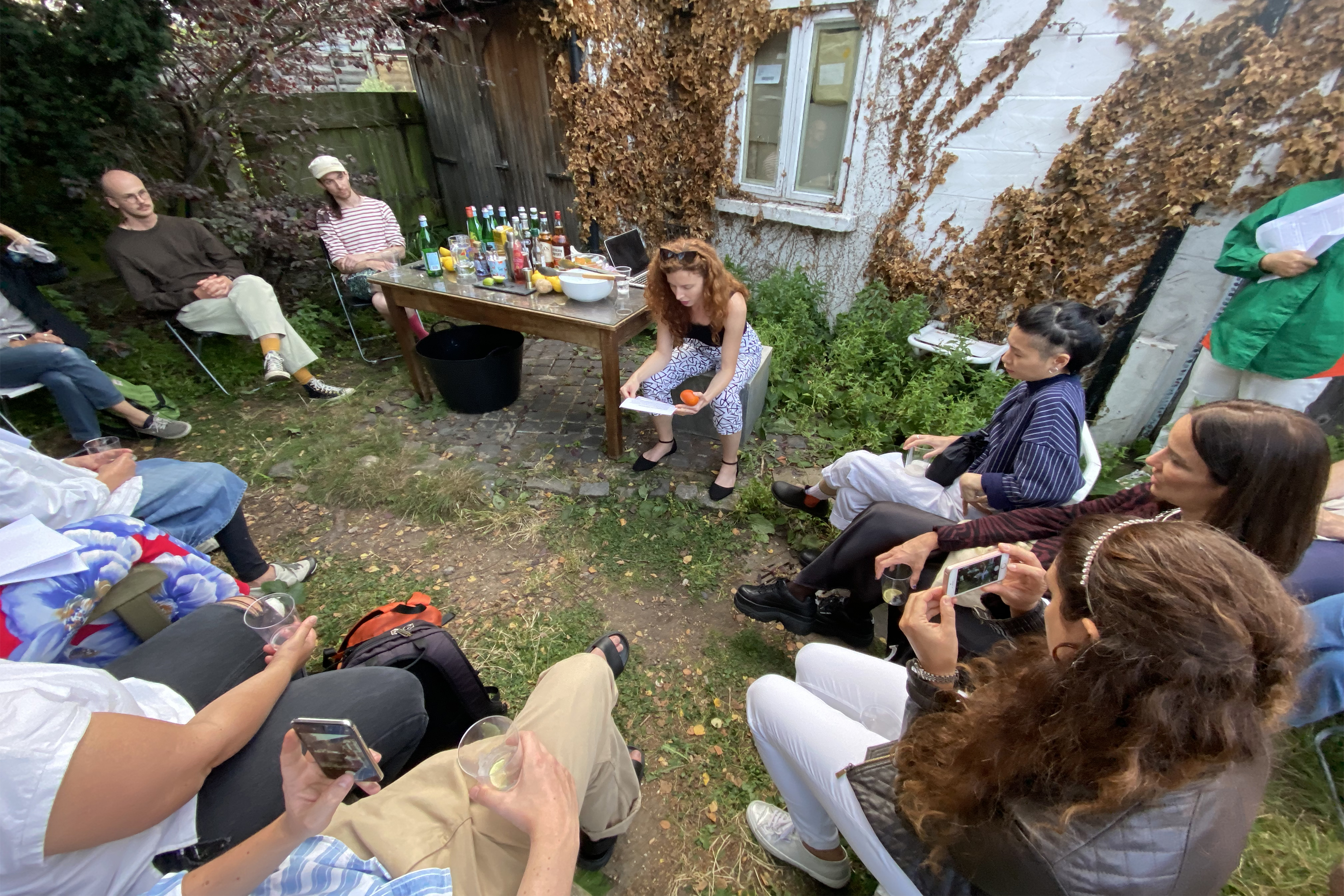 A photograph of people drinking and relaxing sitting on chairs in a semi-circle in a back garden surrounded by greenery.