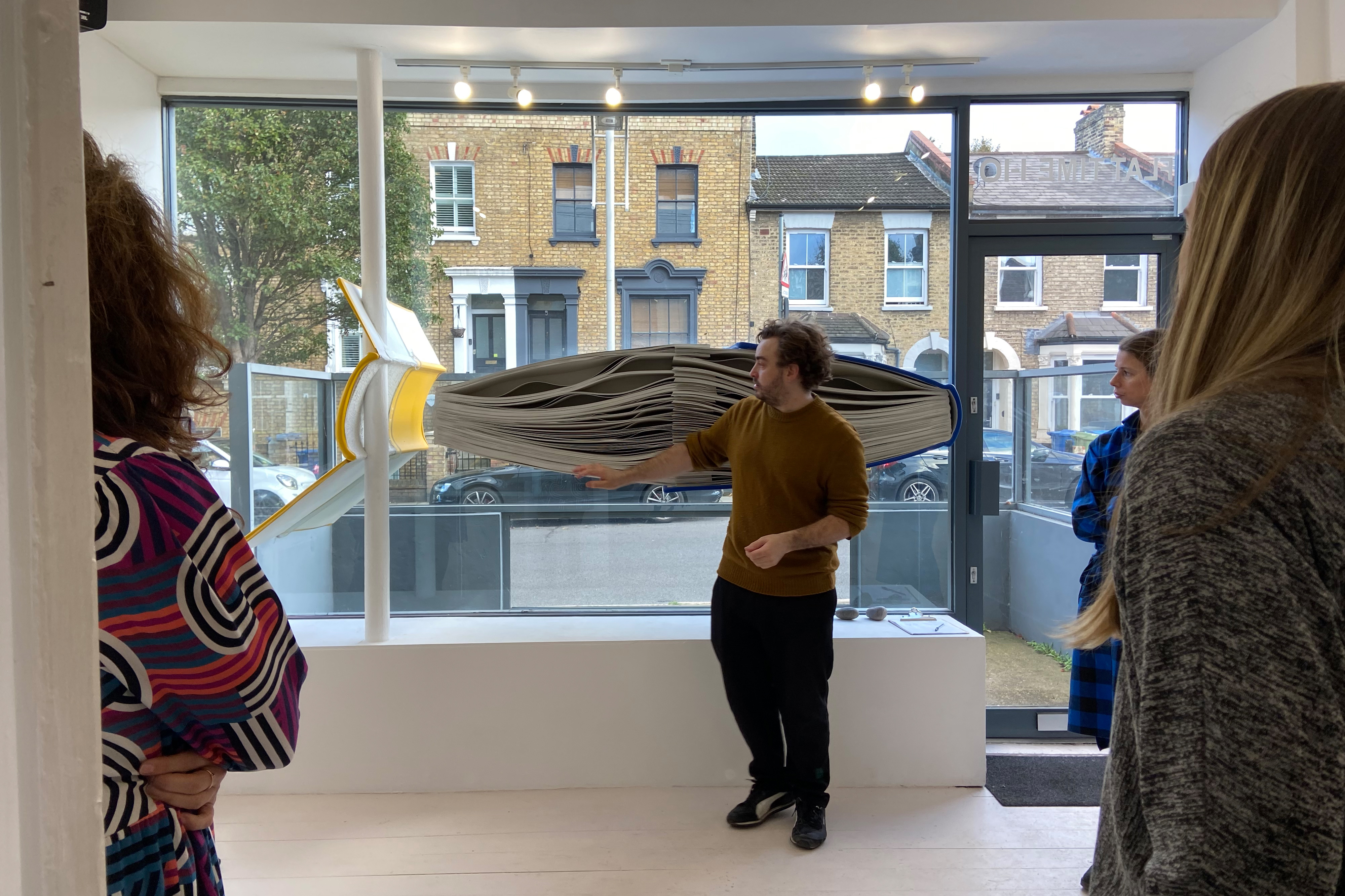 A group of people standing around in a large white room with large windows, watching a man standing next to a large book sculpture that sticks out of the window
