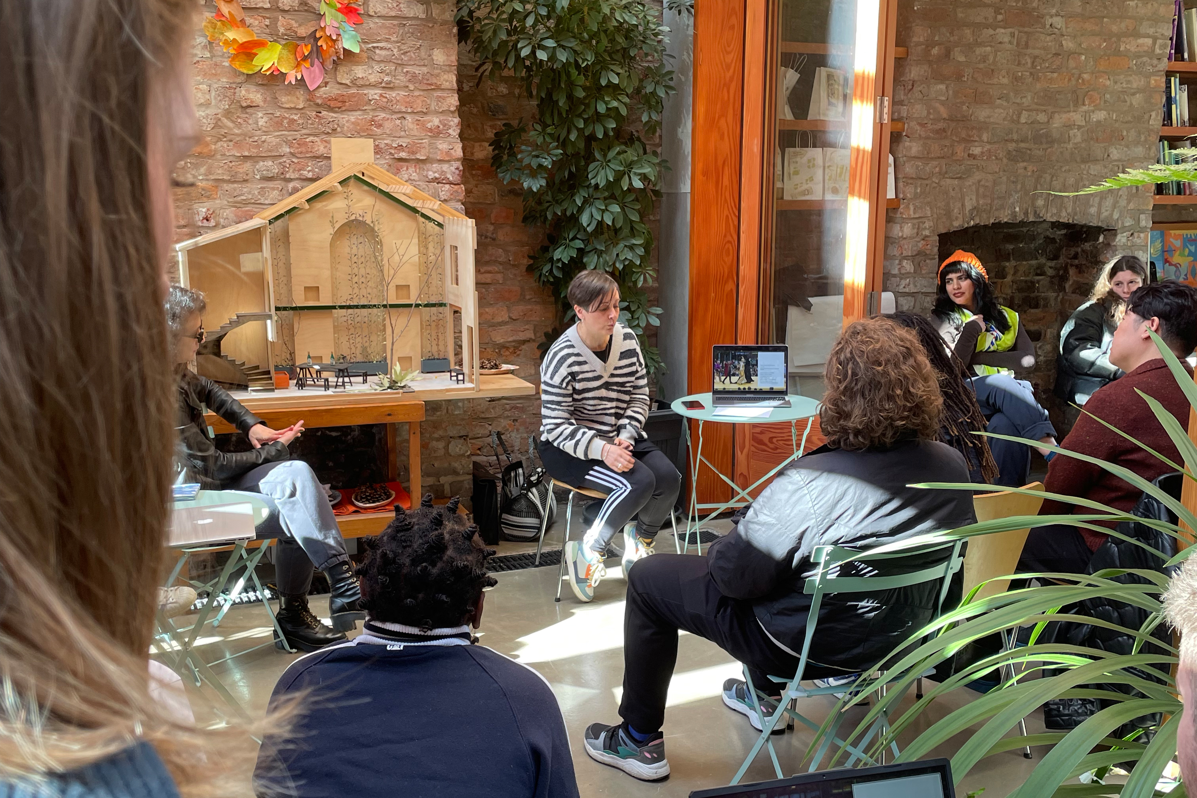 A group of people sitting together in a large open room with grey concrete floor, multiple plants, in front of a large red and grey brick wall