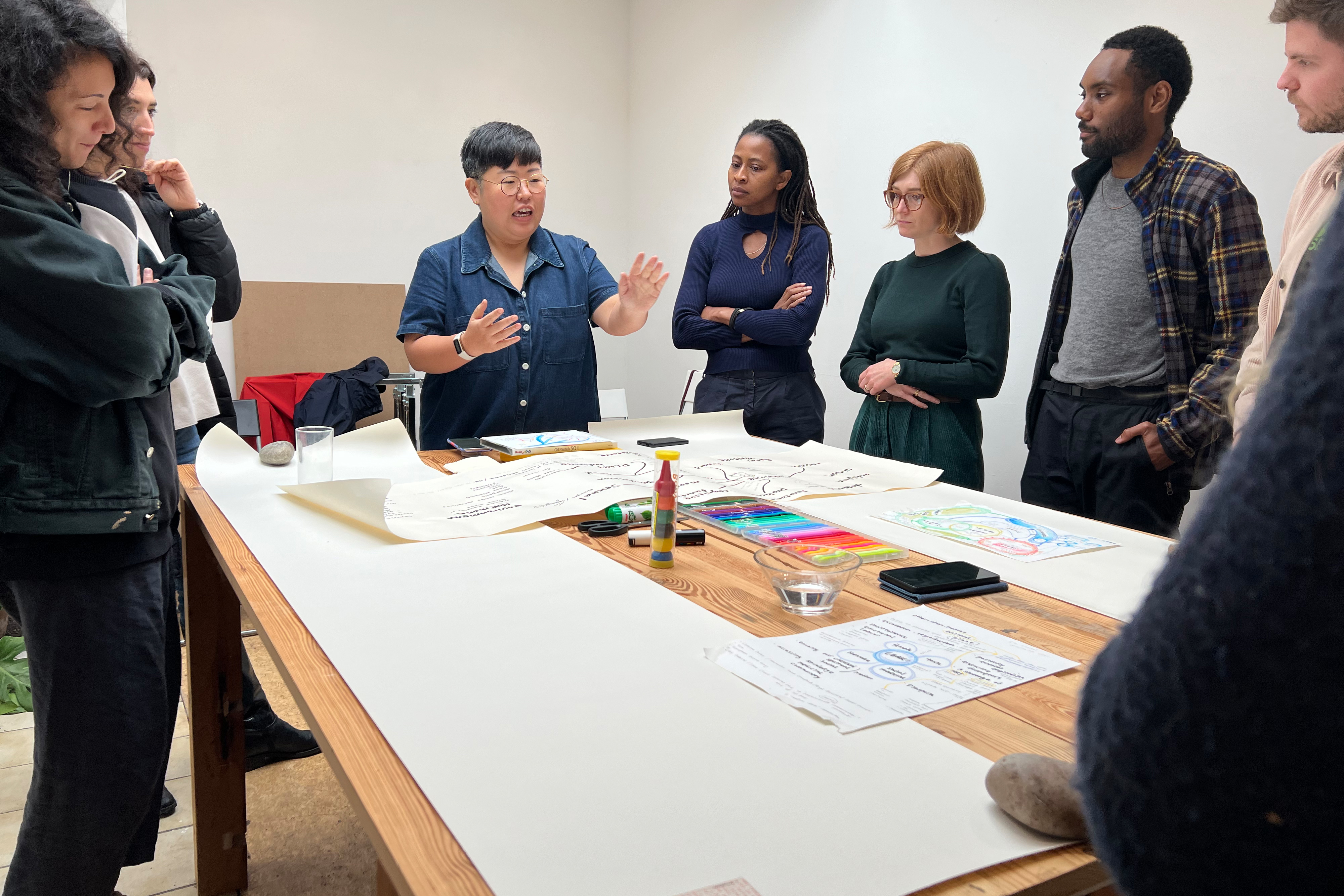 A group of people standing around a large wood table covered in large white rolls of paper and various drawing and painting materials
