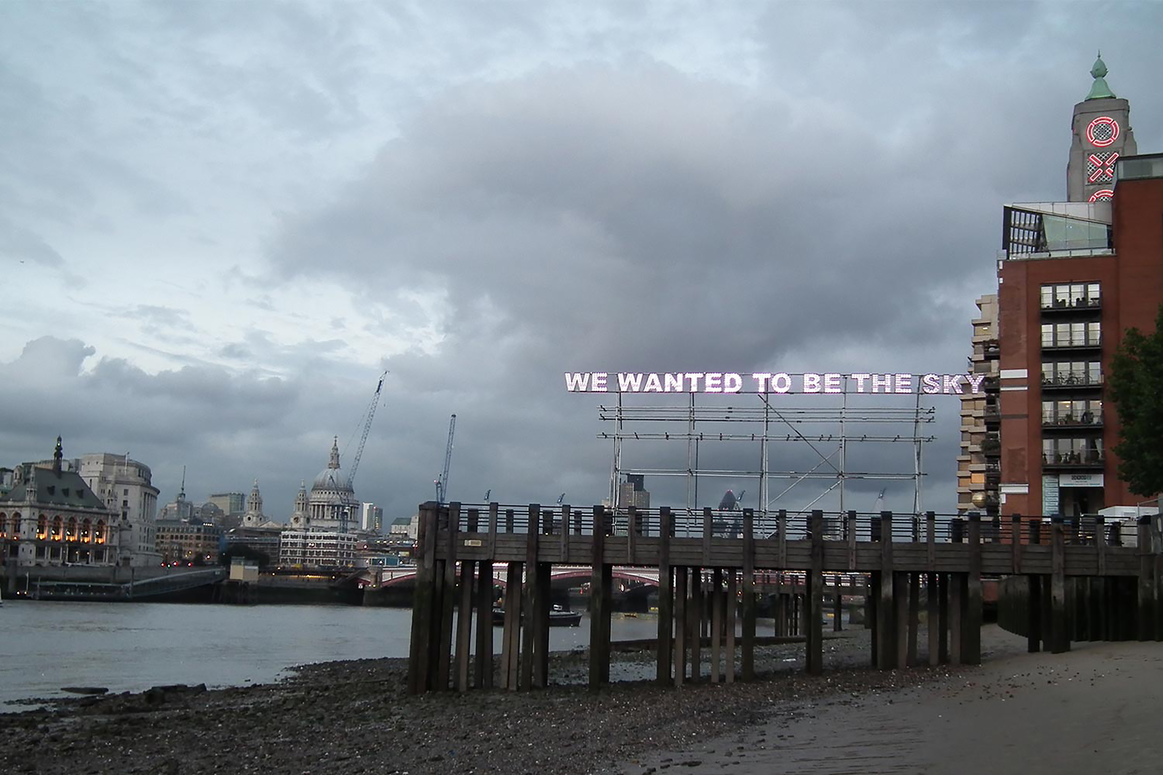 A photograph of a beach with no tide exposing a wooden pier in front of a sign that reads "We Wanted To Be The Sky".