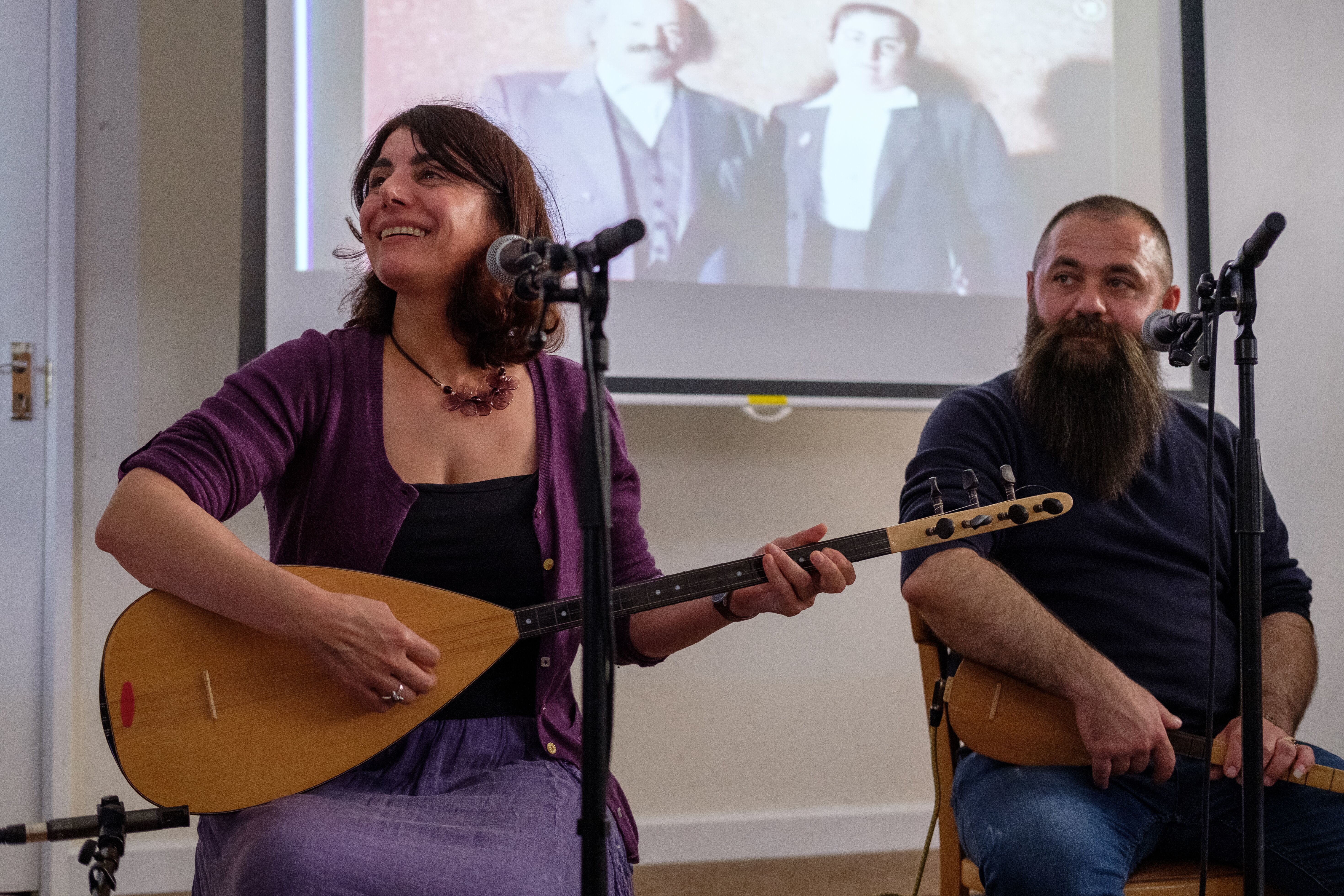Two musicians singing and playing music. Both are sitting behind microphones while one plays a saz. They are in a small room in front of a large presentation screen.
