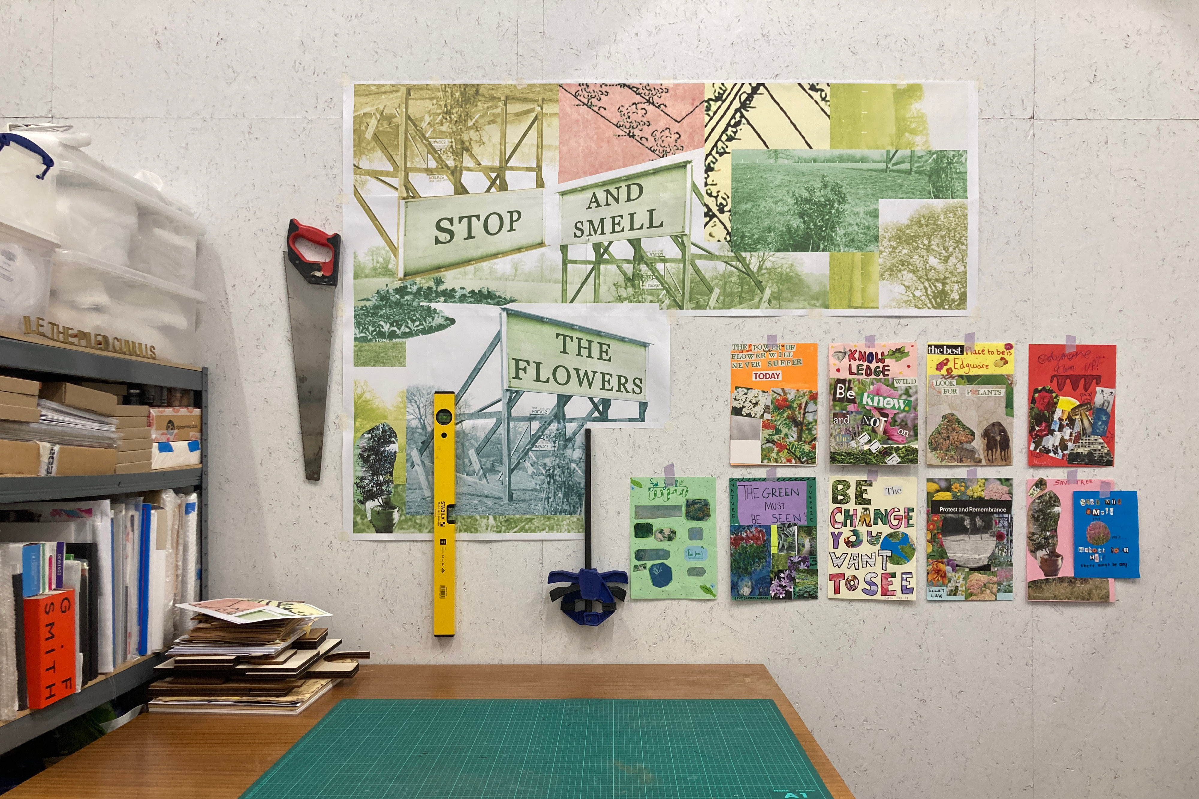 A white wall with a collage of picture and word clippings. The wall is next to a wooden table with a green cutting mat and a shelf full of objects and books.