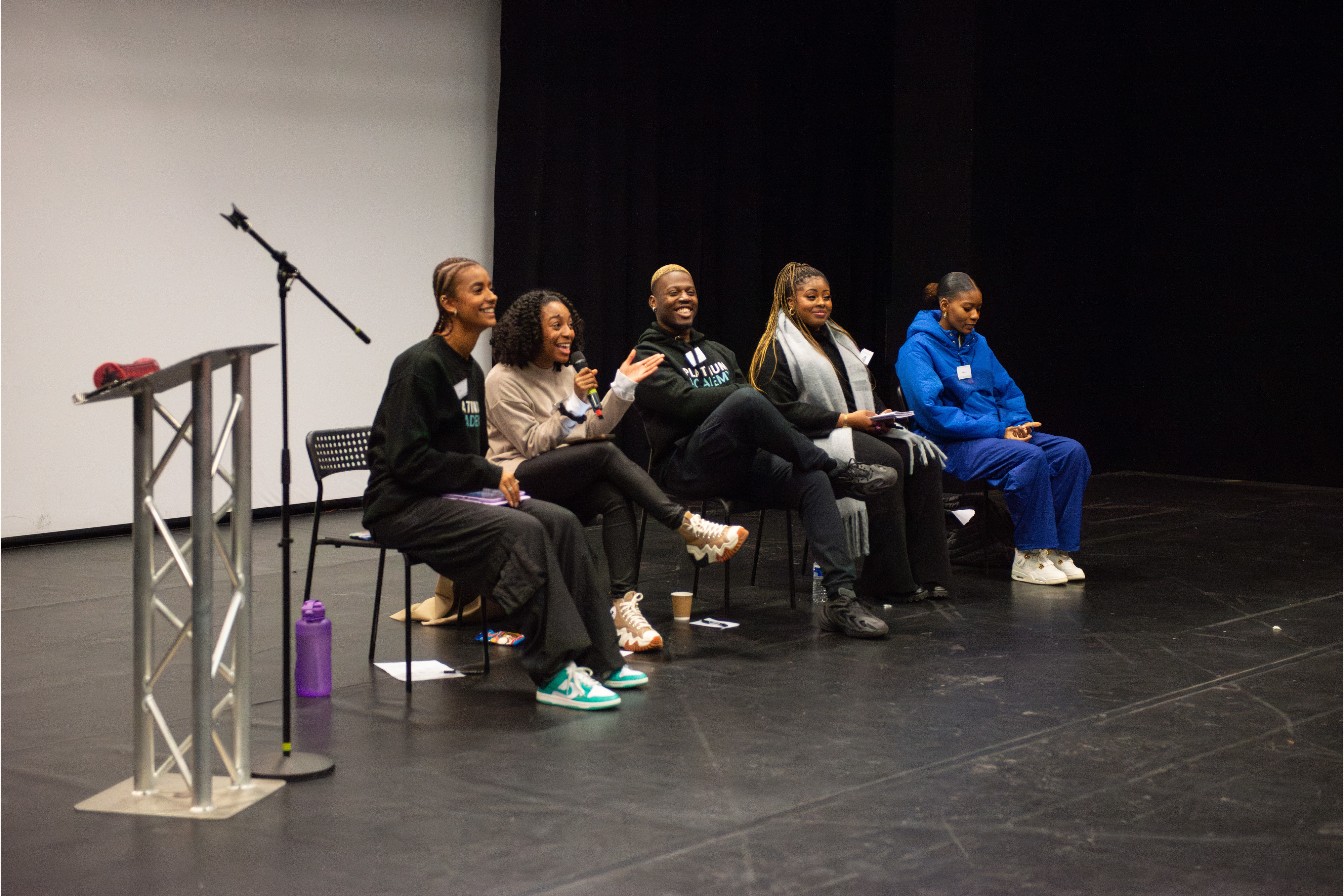 A panel of five people sits on a stage, smiling and speaking to an audience. One person holds a microphone while the others listen attentively. There is a podium and microphone stand in the foreground.
