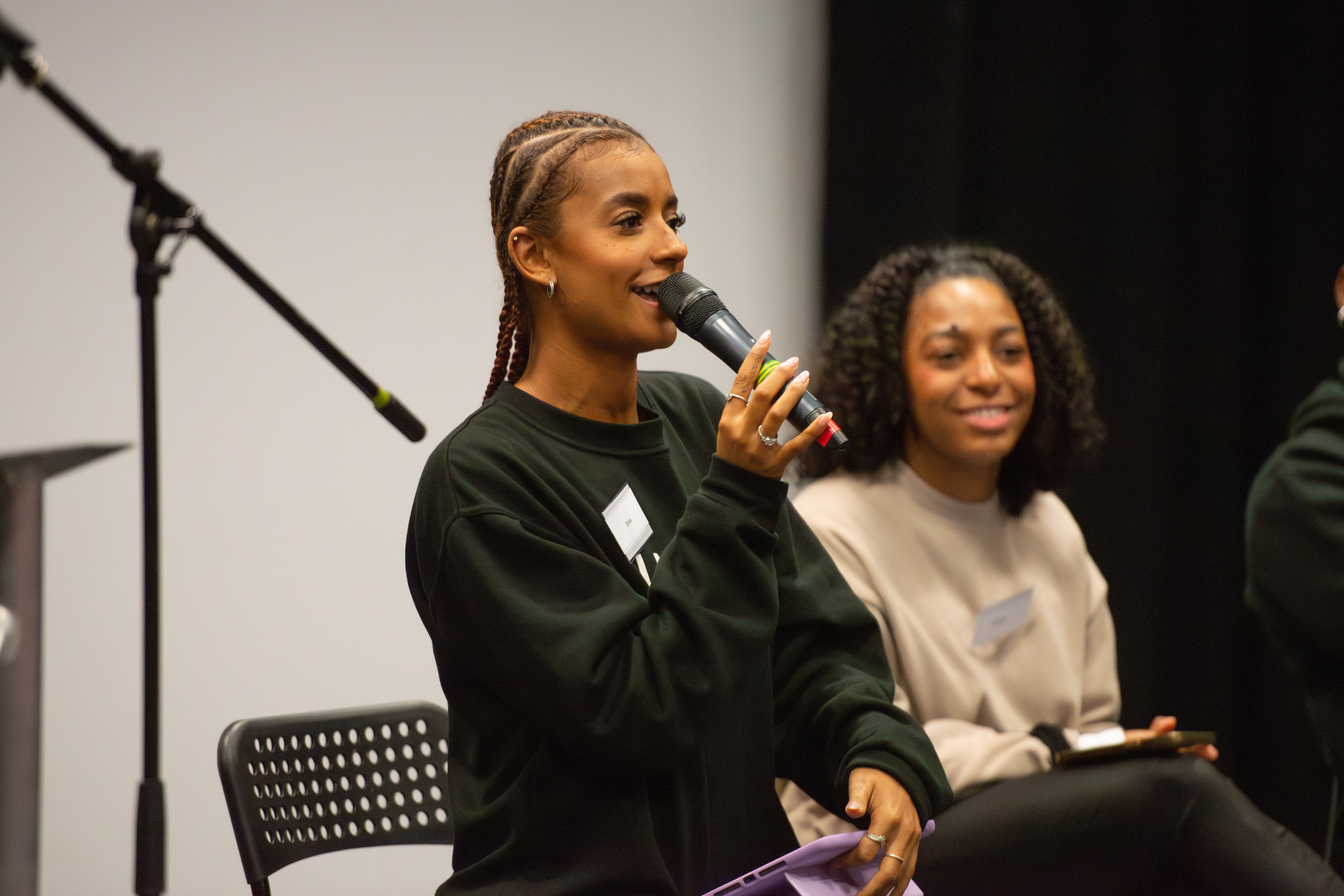 Young woman speaking into a microphone during a panel, with another woman smiling beside her.